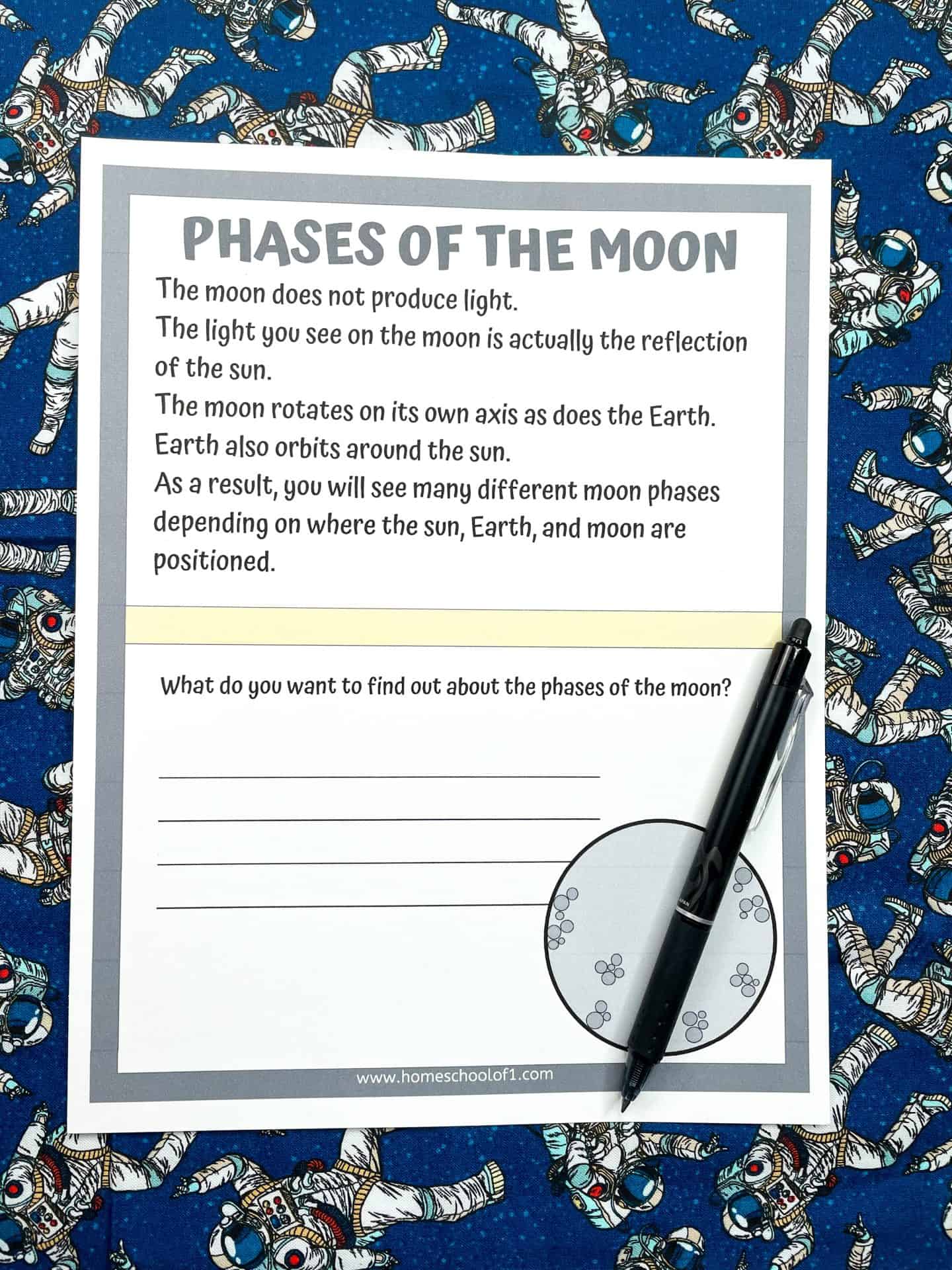 A printed worksheet titled "Phases of the Moon" placed on a blue background with illustrations of astronauts. The worksheet explains that the moon reflects sunlight, rotates on its axis, and appears in different phases due to its position relative to the Earth and sun. It includes a writing prompt asking, "What do you want to find out about the phases of the moon?" with blank lines for student responses. A black pen rests diagonally across a moon phase illustration in the bottom right corner.