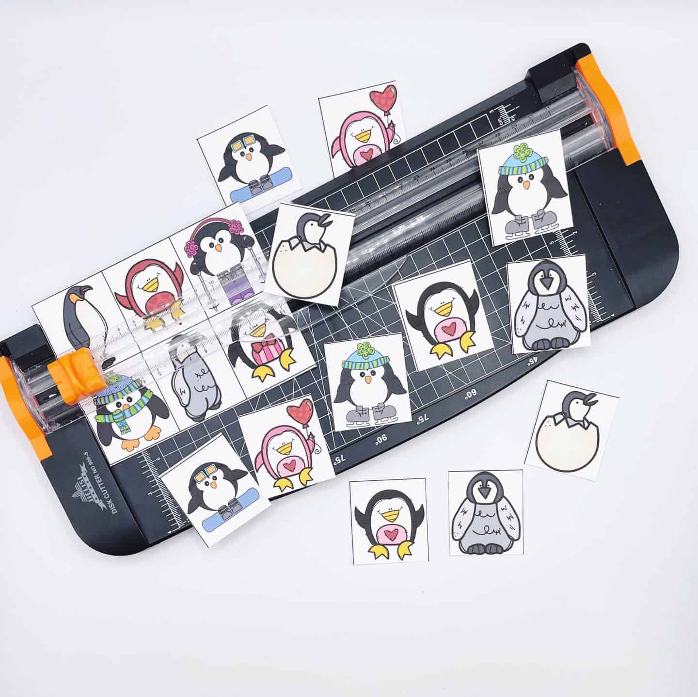 A paper cutter sits on a white surface surrounded by colorful square cards featuring illustrated penguins in different outfits and poses, part of a printable penguin memory game being cut out. Some cards show penguins with accessories like hats, scarves, or holding hearts.

