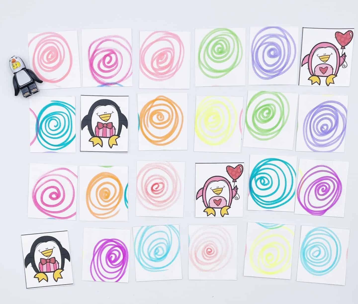 A penguin-themed memory game is laid out on a white surface, featuring square cards with colorful swirly backs. Some cards are flipped to reveal cartoon penguins holding hearts or wearing bow ties. A small penguin figurine is placed to the left of the grid.