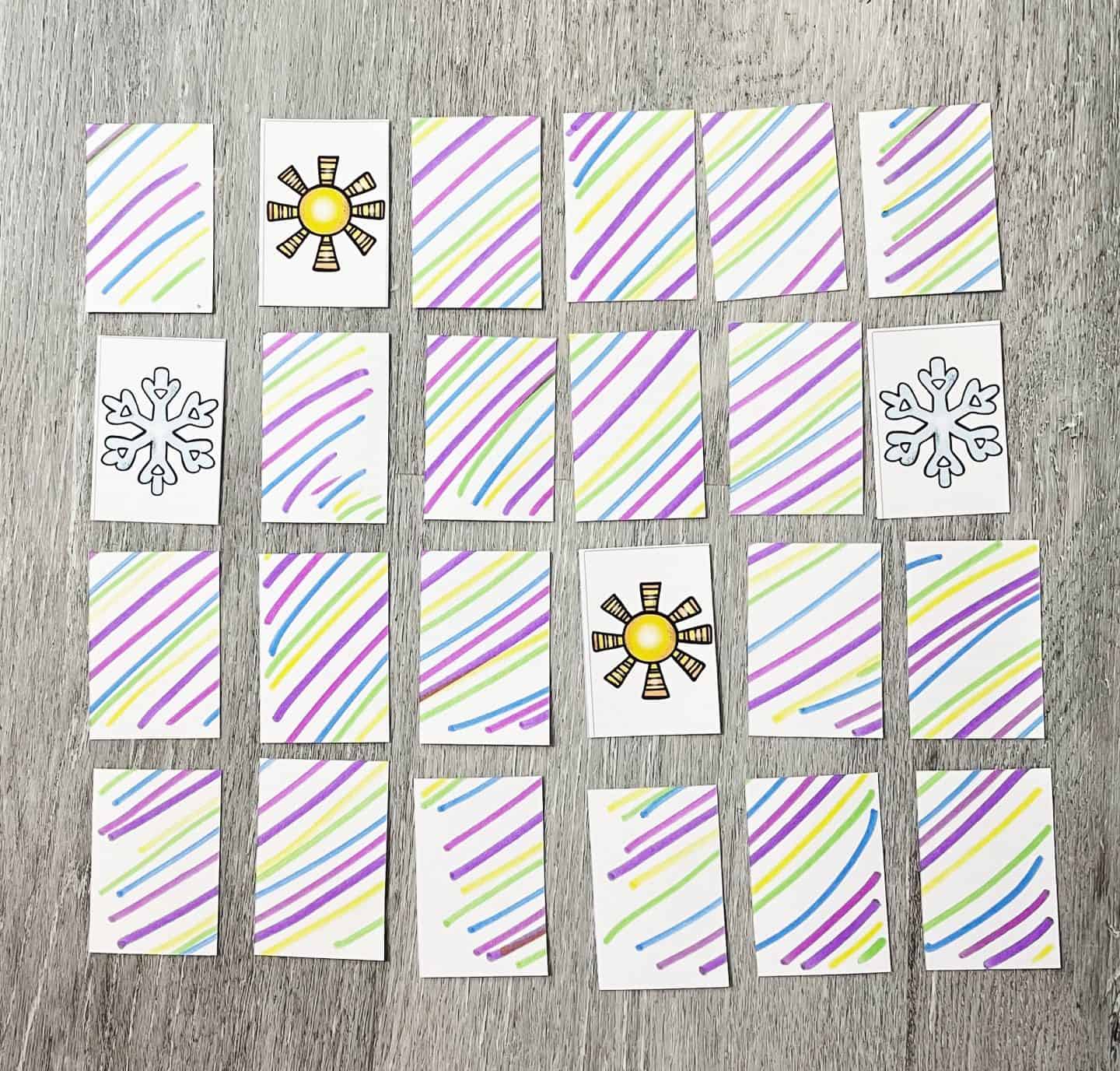 Weather-themed memory game cards laid out face down with a few flipped over, revealing sun and snowflake icons. Each card back is decorated with colorful diagonal stripes in purple, yellow, green, and blue on a wood-textured background.