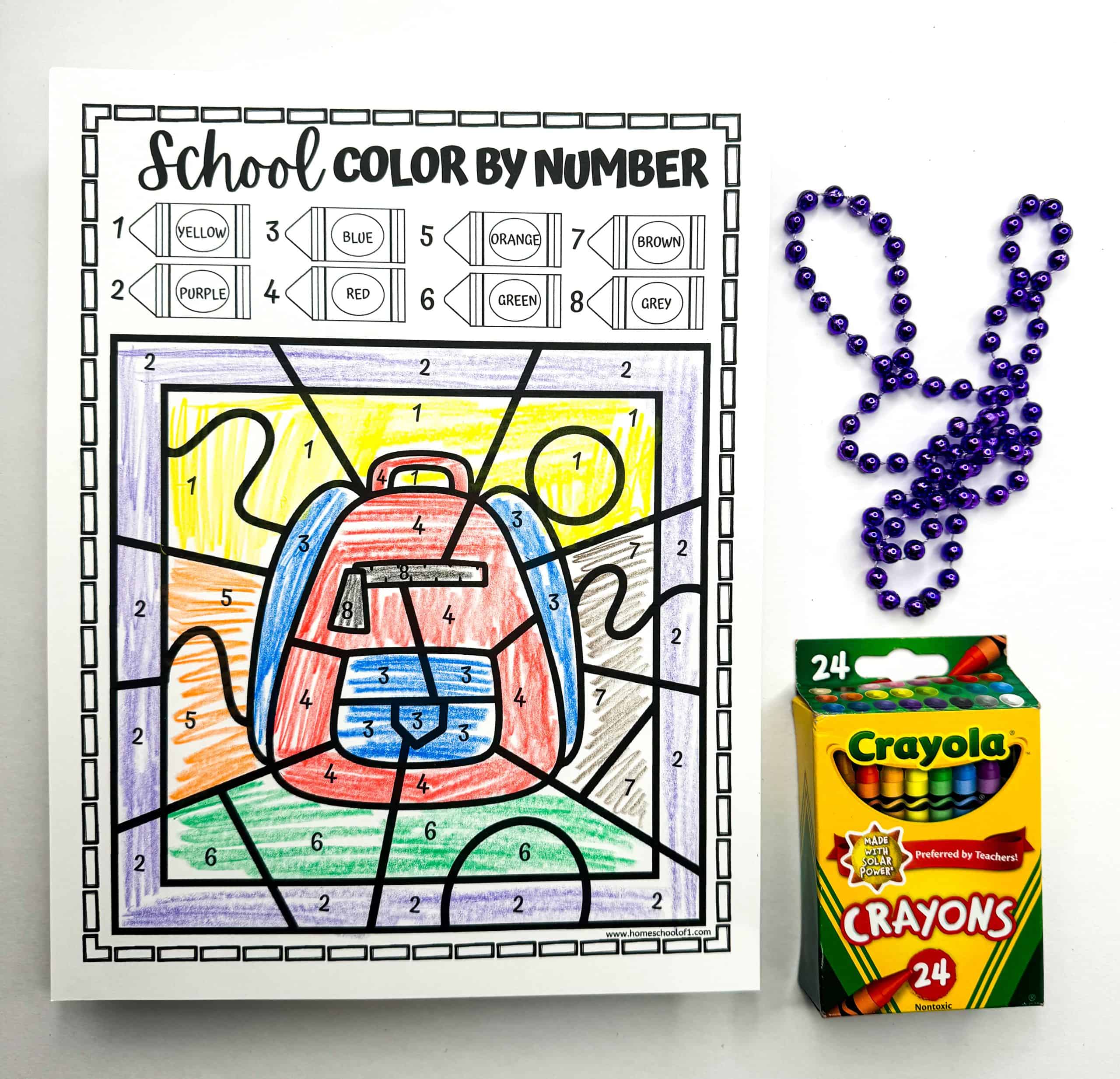 A color by number sheet presents a vibrantly colored school backpack. Alongside the completed worksheet are purple beads arranged as a rabbit and a box of Crayola crayons, highlighting this as a playful learning activity.