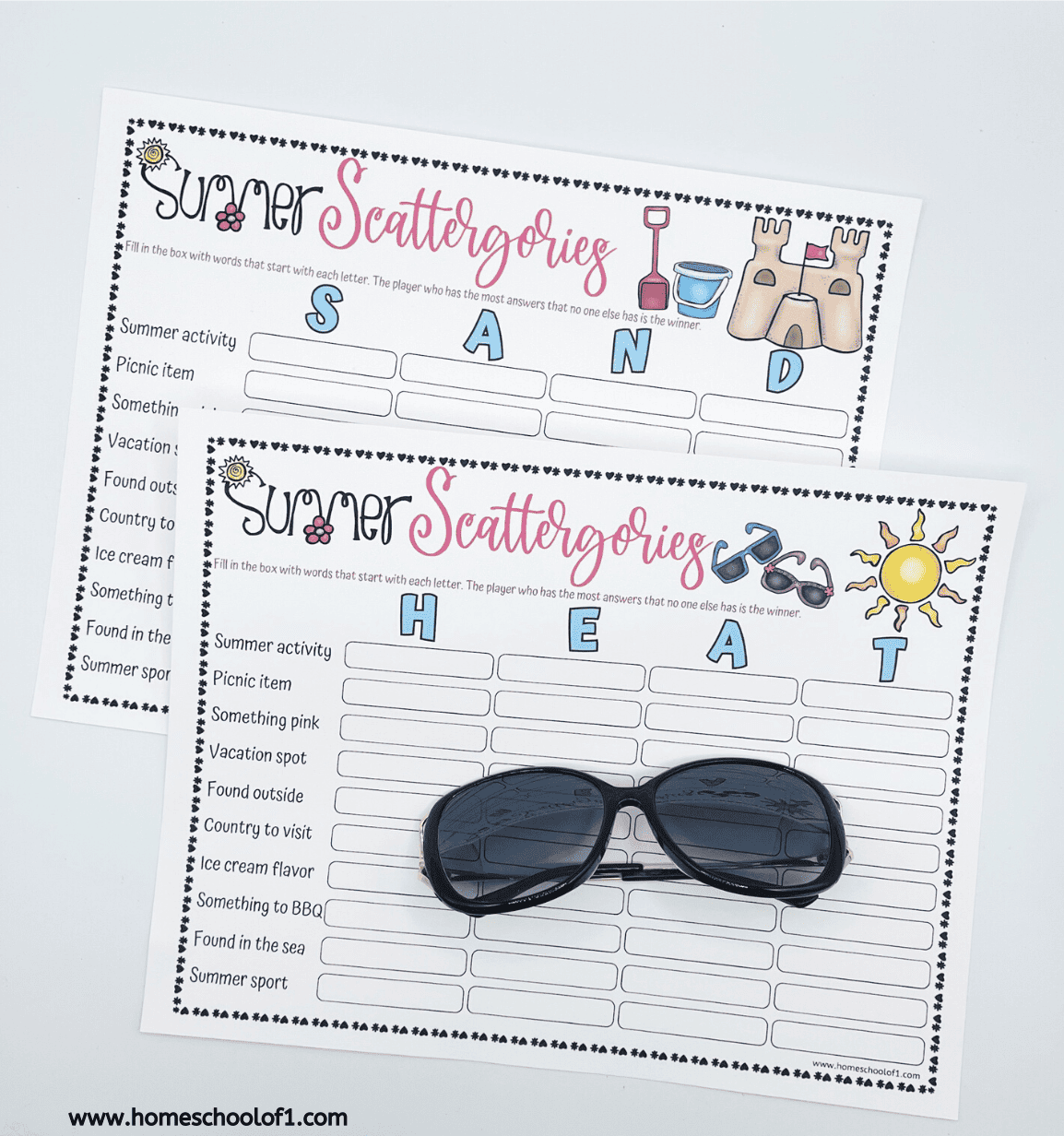 Two blank "Summer Scattergories" game sheets titled "SAND" and "HEAT" are shown, each with themed illustrations—beach toys and a sandcastle for "SAND," and sunglasses with a sun for "HEAT." Categories include summer activity, something pink, country to visit, and more. A pair of black sunglasses rests on the "HEAT" sheet, all placed on a white surface.