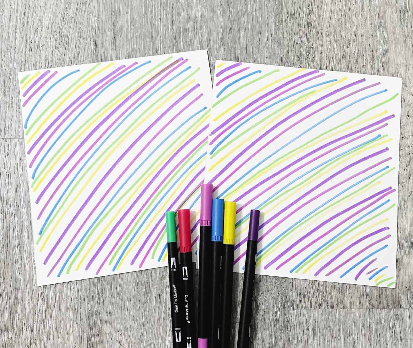 Two sheets of white paper with hand-drawn colorful diagonal stripes in purple, green, yellow, and blue, placed on a wooden surface. A set of six dual tip markers in coordinating colors is arranged below the sheets.