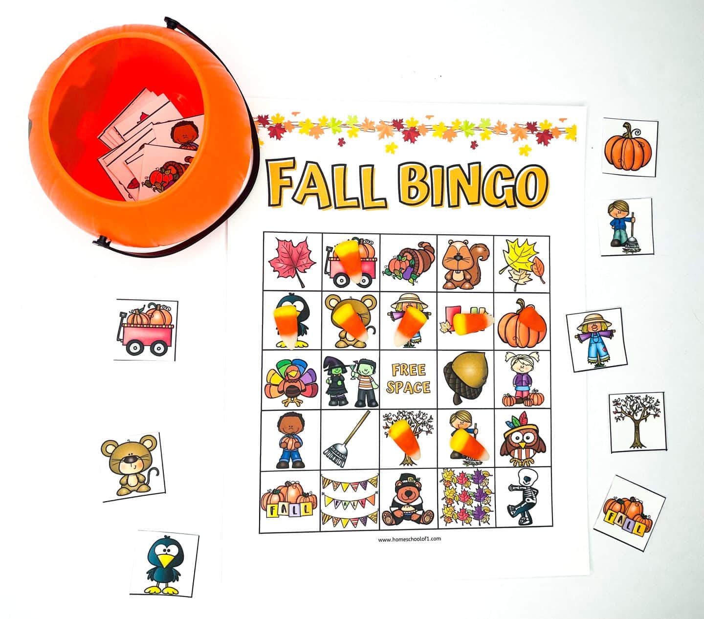 A festive Fall Bingo printable game setup with an autumn-themed bingo card featuring colorful icons like pumpkins, scarecrows, acorns, and leaves. Candy corn pieces are used as markers, and bingo calling cards are stored in an orange pumpkin-shaped container. This Fall Bingo printable is perfect for seasonal classroom activities, family fun, or homeschool celebrations.