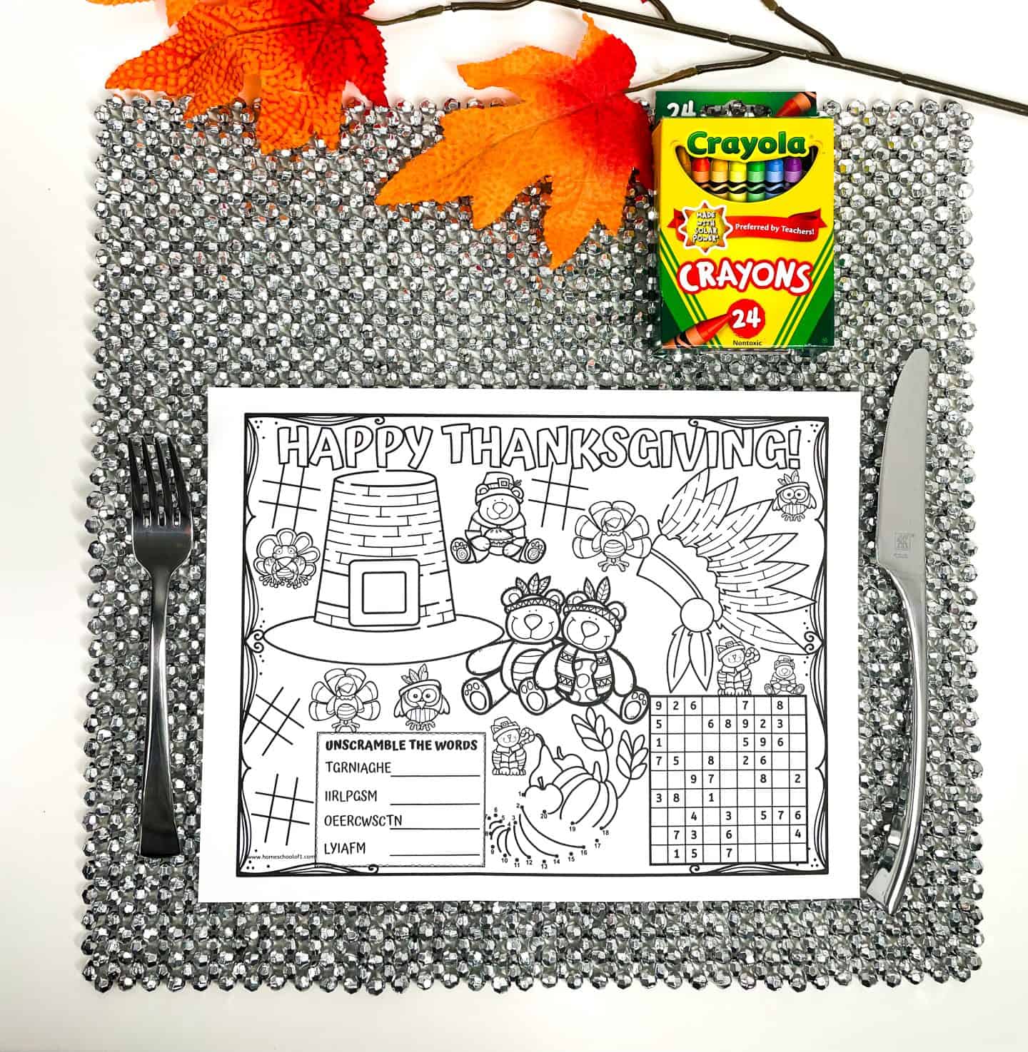 Thanksgiving-themed kids’ placemat on a sparkly silver mat with a fork and knife, featuring puzzles and coloring activities like word scrambles, dot-to-dot, and a number grid. The sheet reads “Happy Thanksgiving” and includes illustrations of pumpkins, Pilgrims, and fall symbols. A box of Crayola crayons and orange fall leaves rest at the top.