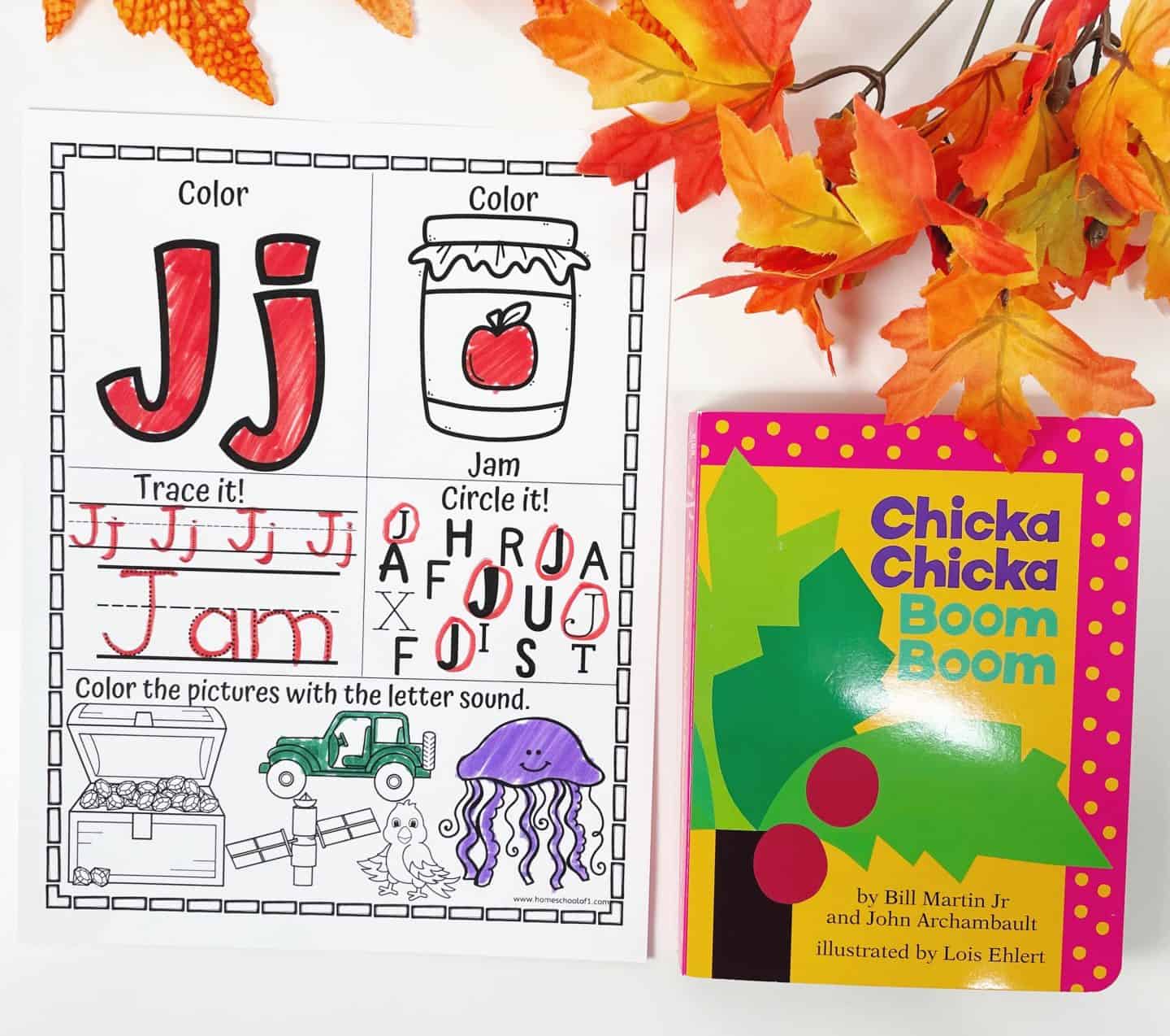 A Thanksgiving-themed alphabet worksheet highlights the letter J. The uppercase and lowercase "Jj" are colored red, with a jar of jam illustration next to them. The word "Jam" is traced, and students circle all the J’s from a mixed group of letters. At the bottom, images like a jellyfish are colored to match the letter sound. The worksheet is paired with the book Chicka Chicka Boom Boom, placed on the right, and decorated with colorful autumn leaves.