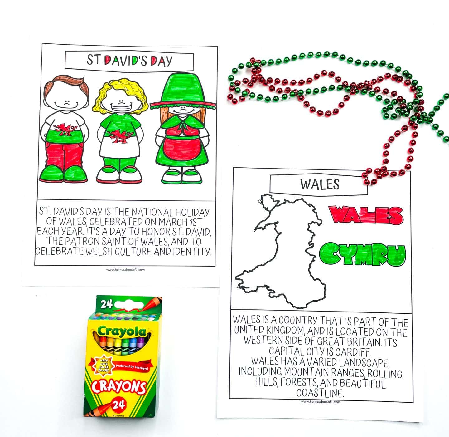 Two completed Welsh-themed coloring pages are shown. One celebrates St. David’s Day with three cartoon children dressed in red, green, and white, alongside a description of the March 1st holiday. The second page outlines the shape of Wales with the words “Wales” and “Cymru” in red and green, plus a short paragraph describing the country’s geography and capital, Cardiff. Red and green bead necklaces and a box of Crayola crayons are also included in the scene.