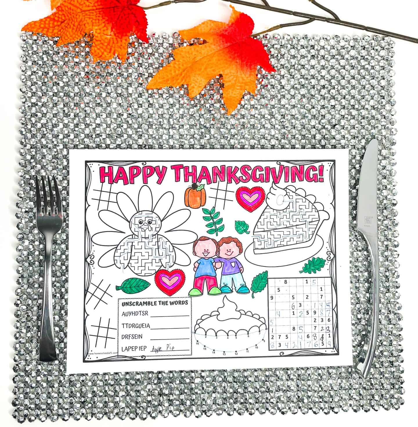 Colorful Thanksgiving placemat set on a silver beaded charger with a fork on the left and knife on the right. The placemat includes mazes, tic-tac-toe, a dot-to-dot pie, a number puzzle, and a word scramble with answers partially filled in. It features festive illustrations like a turkey, pie, kids, and fall leaves, with some areas colored in pink, blue, green, and orange. Fall leaves decorate the top.