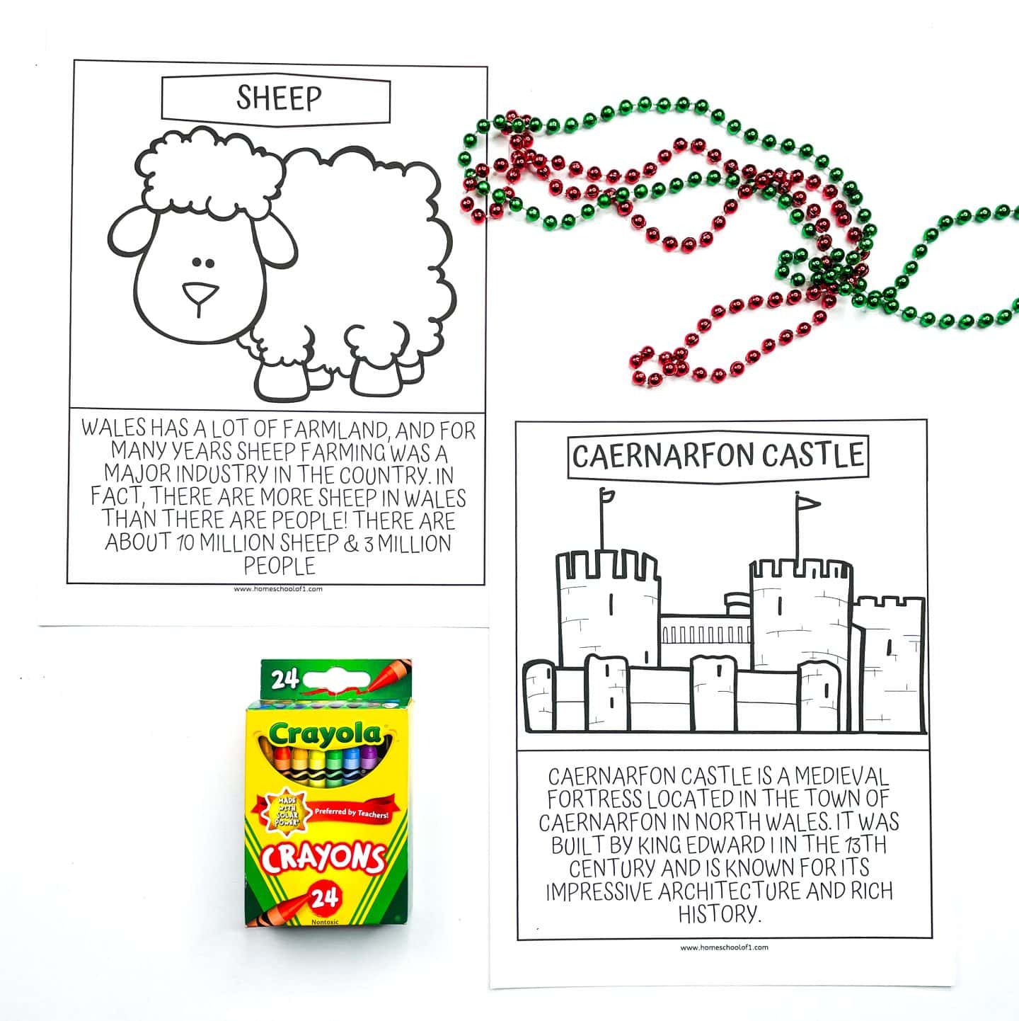 Two uncolored Welsh-themed educational coloring pages are displayed. One features a cartoon sheep and a paragraph explaining that Wales has more sheep than people—about 10 million sheep to 3 million people. The second shows Caernarfon Castle with text describing it as a medieval fortress built by King Edward I in the 13th century. The setup includes a box of Crayola crayons and a festive strand of red and green beads.