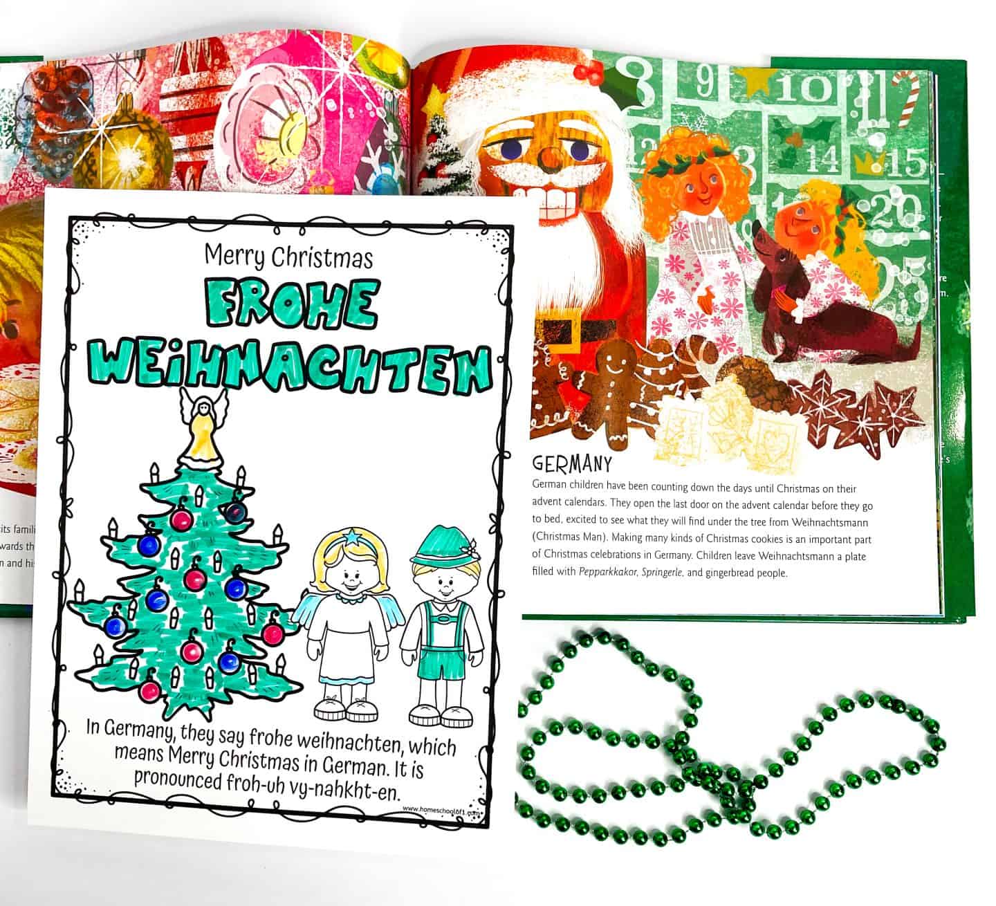 Coloring page featuring a German Christmas greeting with the words "Merry Christmas Frohe Weihnachten" in large green letters. Below, a decorated Christmas tree stands between a girl dressed as an angel and a boy in traditional German clothing. The text explains how to say and pronounce “Frohe Weihnachten.” Behind the worksheet is an open book showing a festive scene with Santa, children, Christmas cookies, and an advent calendar. A strand of green beads lies nearby.