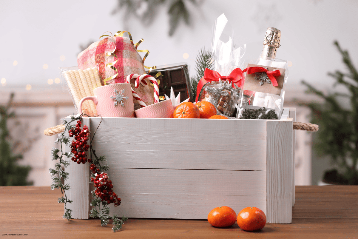 A white wooden Christmas Eve box filled with festive items including mugs, wrapped gifts, chocolate, biscuits, a small bottle of alcohol, clementines, and holiday greenery, set on a wooden table with a softly lit, decorated background.