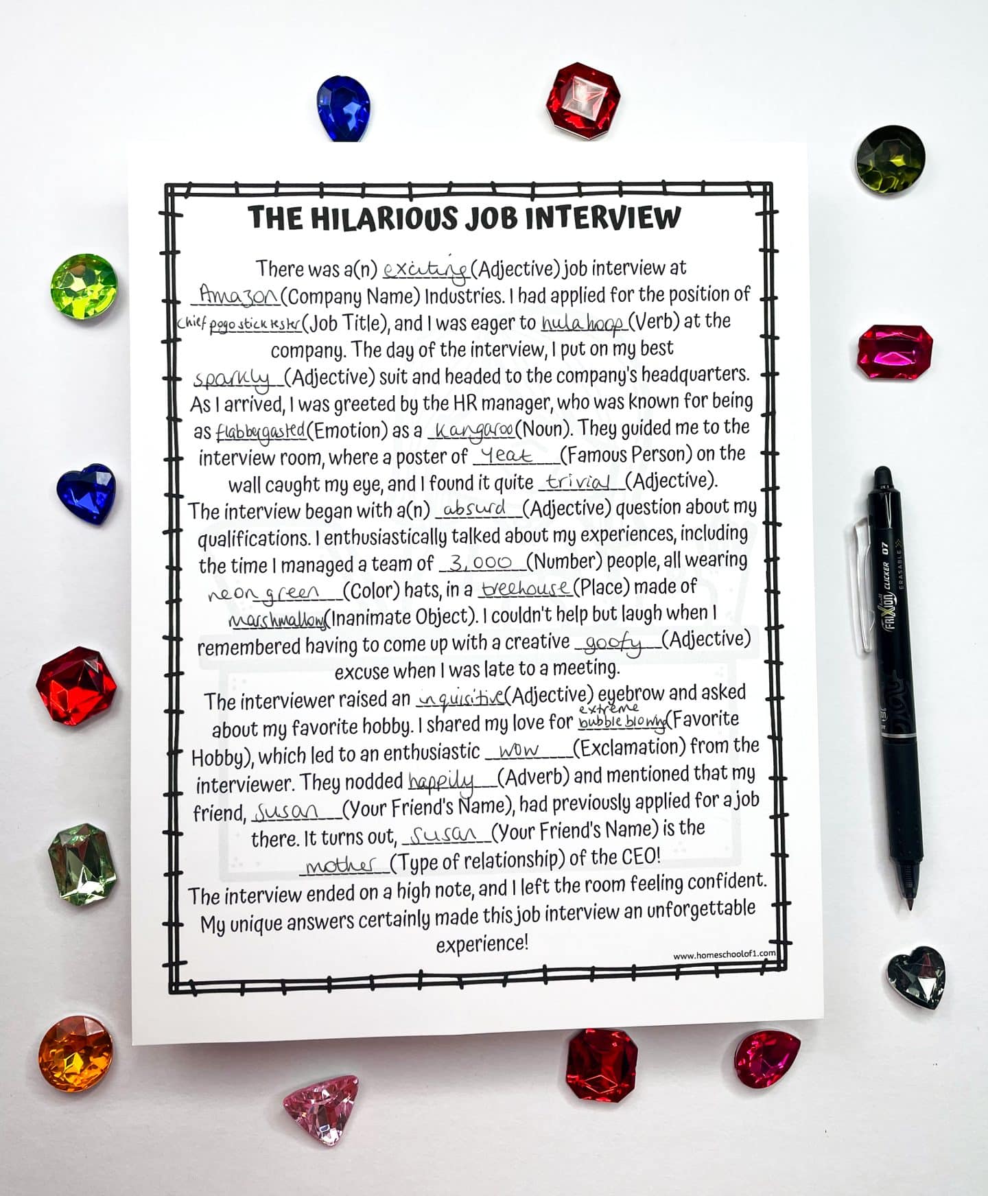 Completed Mad Libs worksheet titled “The Hilarious Job Interview,” featuring wacky answers like “hula hoop,” “kangaroo,” “bubble blowing,” and “Susan.” The story humorously describes a bizarre job interview experience with creative adjectives and nouns. The page is surrounded by colorful plastic gems and a black pen.