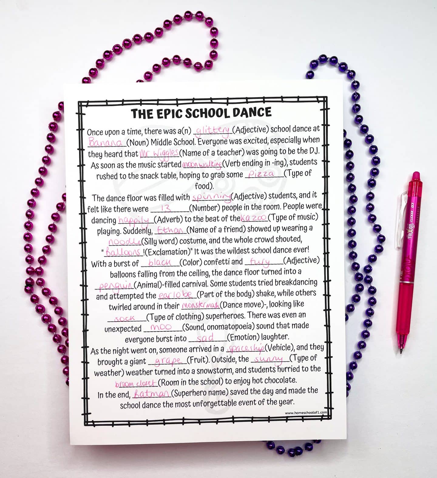 Completed Mad Libs worksheet titled “The Epic School Dance,” filled with funny words like “banana,” “moonwalking,” “noodle,” and “batman.” The story humorously describes a chaotic school dance featuring kazoo music, penguins, and a sock-wearing superhero. The page is surrounded by pink and purple beaded necklaces, with a pink pen on the right side.