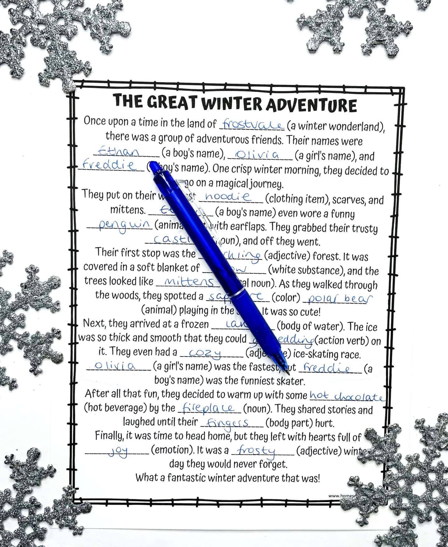 A completed winter-themed Mad Libs worksheet titled “The Great Winter Adventure” is shown with handwritten answers in blue ink. The story includes playful winter-themed words like “frostvale,” “hoodie,” “penguin,” and “hot chocolate.” A blue pen lies diagonally across the page, which is framed by a black border and decorated with silver glitter snowflakes in the corners.