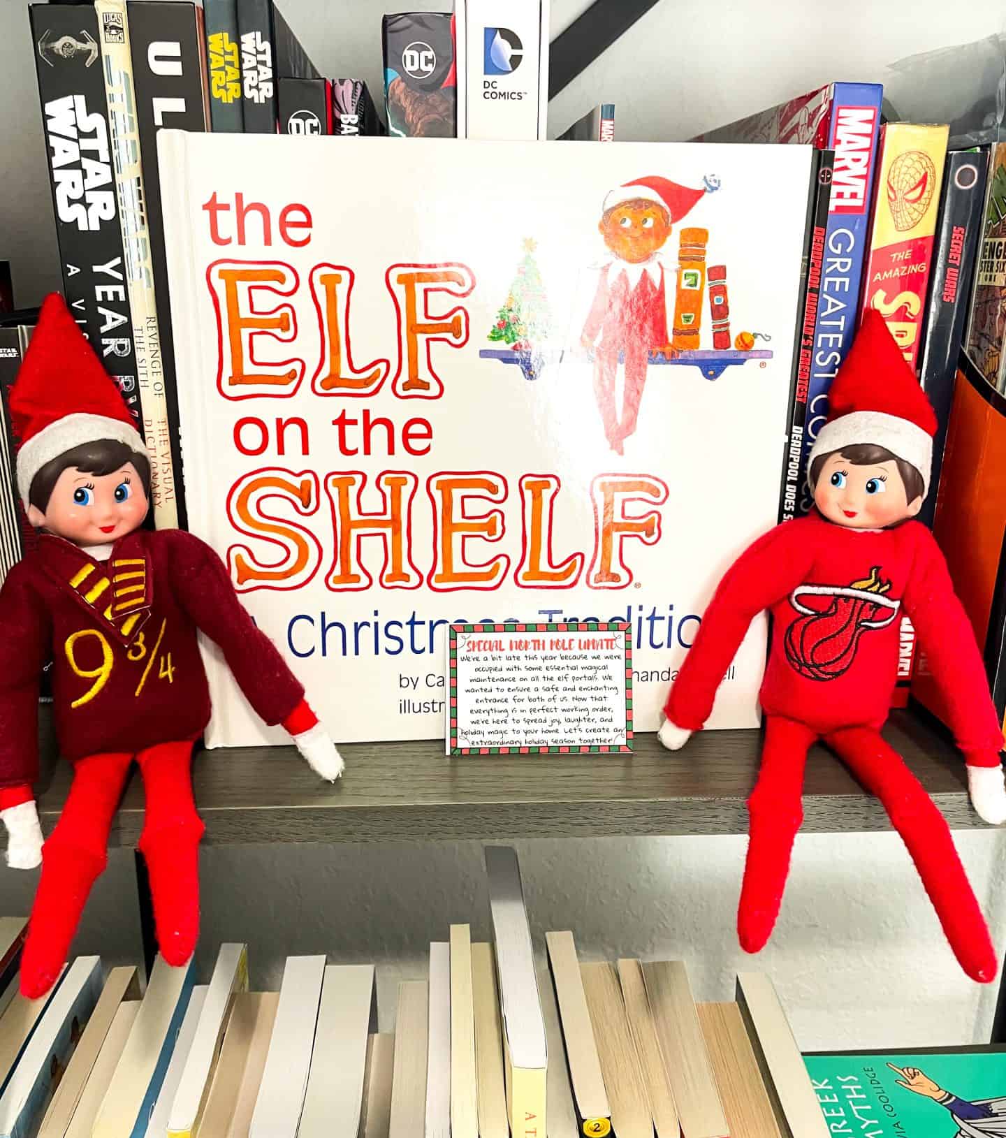 Two Elf on the Shelf dolls sit on a bookshelf on either side of the book The Elf on the Shelf: A Christmas Tradition, surrounded by graphic novels like Star Wars and Marvel comics. One elf wears a Harry Potter-themed outfit with a maroon sweater reading “9¾,” while the other wears a red outfit with a Miami Heat logo. A “Special North Pole Update” letter is placed in front of the book.