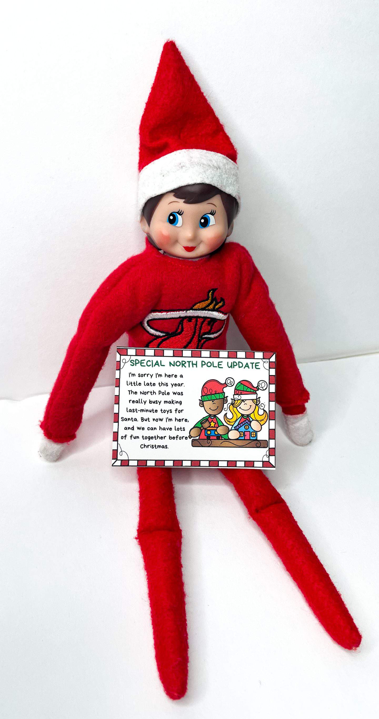 An Elf on the Shelf doll dressed in red with a white-trimmed hat sits against a white background, holding a "Special North Pole Update" letter. The note explains the elf’s late arrival due to busy toy-making at the North Pole, with a cheerful illustration of two cartoon elves working.
