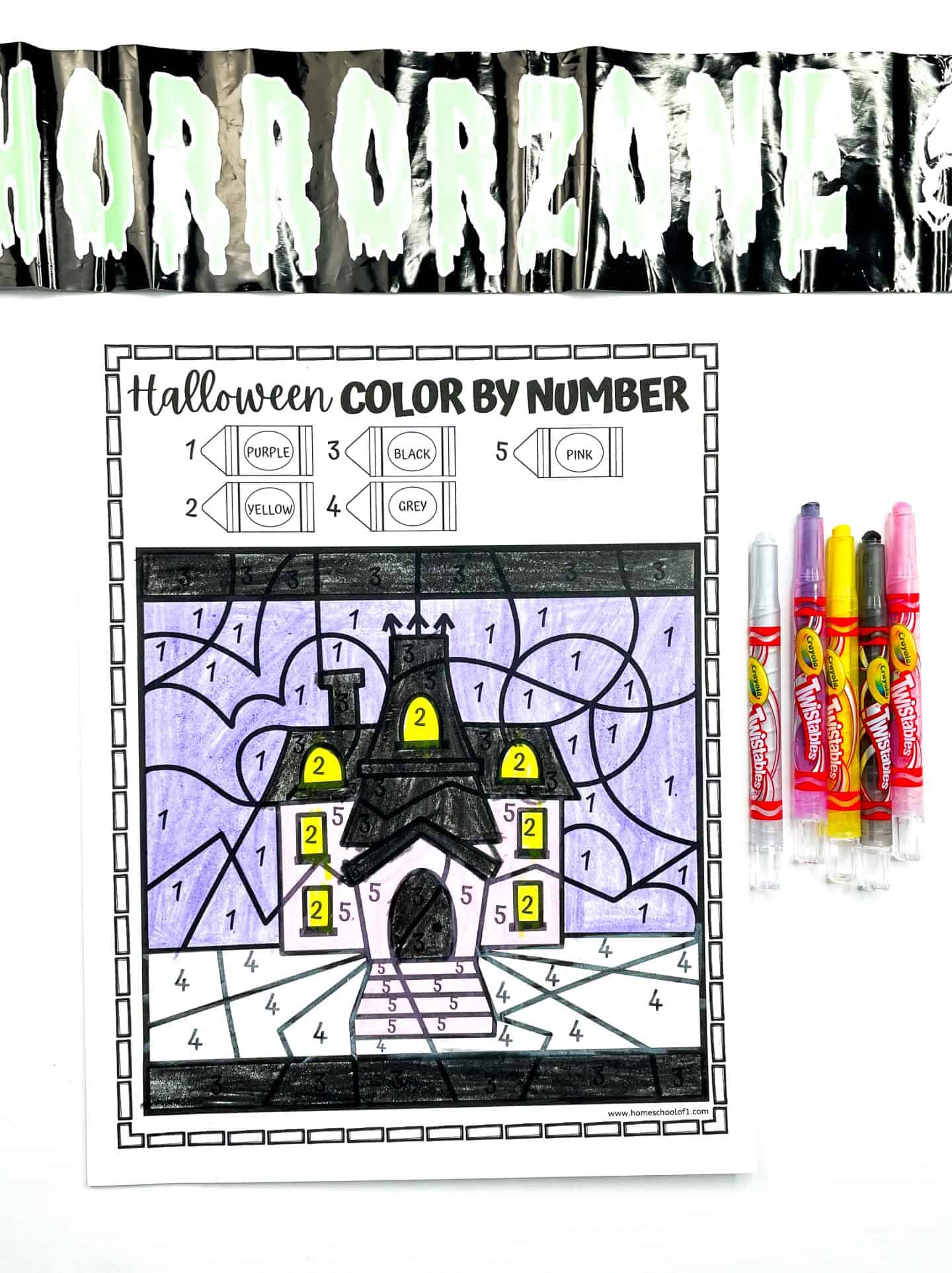  A completed Halloween color-by-number worksheet shows a haunted house with a purple sky, black roof, yellow windows, and grey ground. The color key at the top includes purple, yellow, black, grey, and pink. The page is surrounded by Halloween-themed items including Crayola Twistables markers on the right and black caution tape labeled “HORRORZONE” with dripping white letters across the top.
