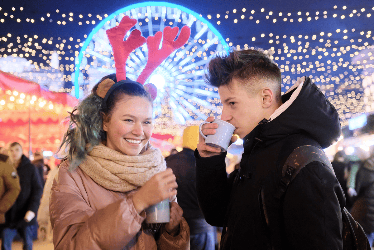 Two teenagers enjoying hot drinks at a festive holiday market at night, with twinkling lights, a Ferris wheel, and a cheerful atmosphere in the background. One teen wears reindeer antlers and smiles, while the other sips from a mug.