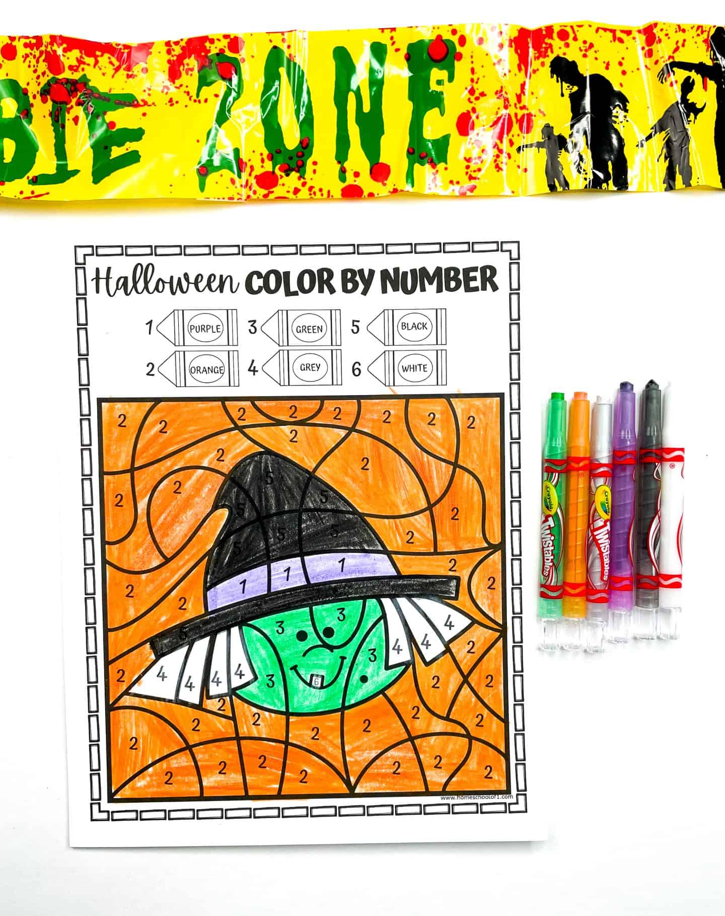A completed Halloween color-by-number worksheet features a friendly green-faced witch wearing a black hat and surrounded by an orange background. The coloring guide at the top includes colors like purple, orange, green, grey, black, and white. To the right are six Crayola markers, and above the page is a yellow “Zombie Zone” caution tape splattered with red and decorated with silhouettes of zombies. 