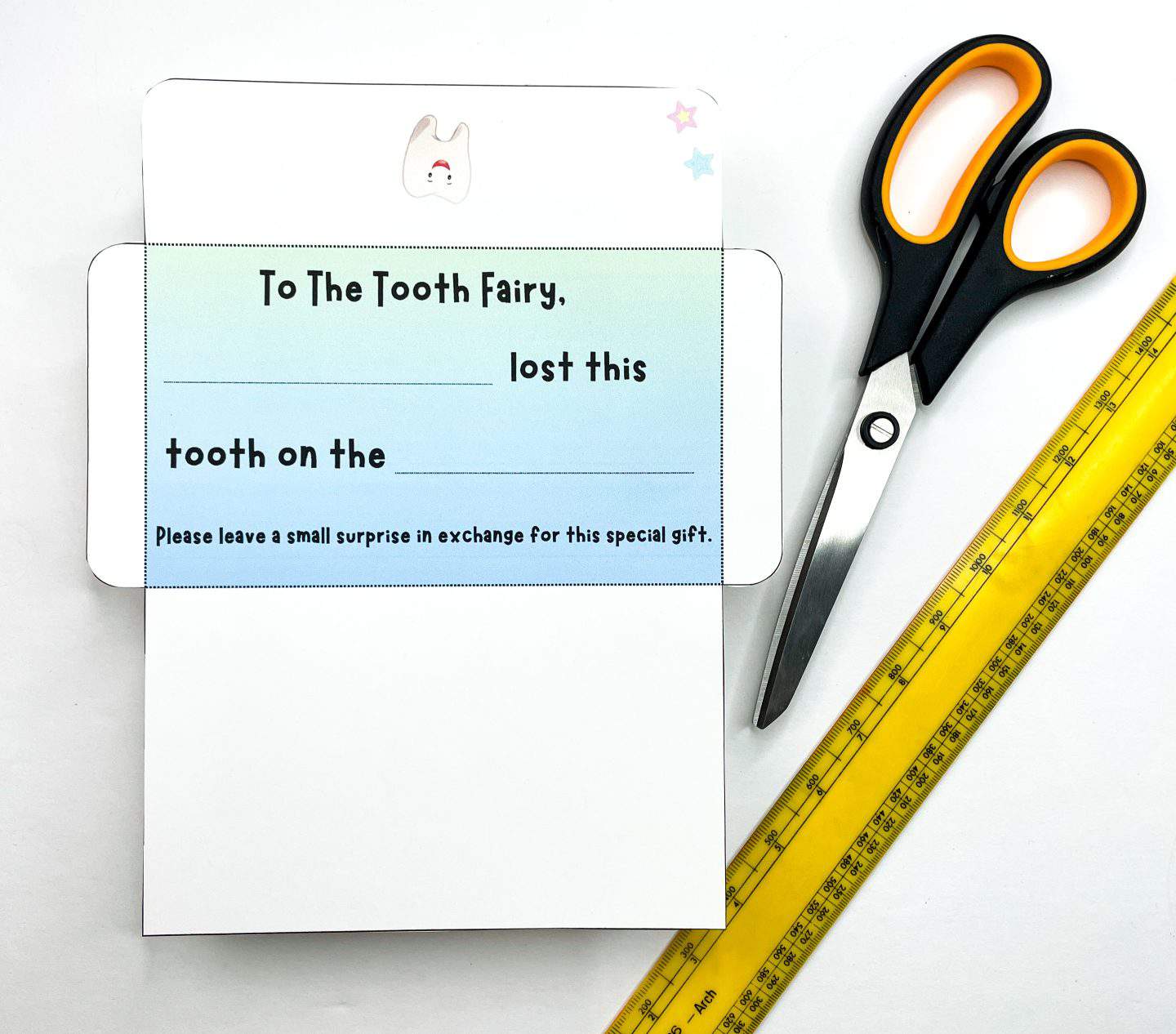 This image shows a partially cut and folded DIY Tooth Fairy envelope placed next to a pair of black and yellow scissors and a yellow ruler. The envelope features a smiling tooth graphic at the top, pastel blue gradient background, and text fields that read: “To The Tooth Fairy, _______ lost this tooth on the _______. Please leave a small surprise in exchange for this special gift.” The layout and tools suggest this is a kids' craft project in progress.