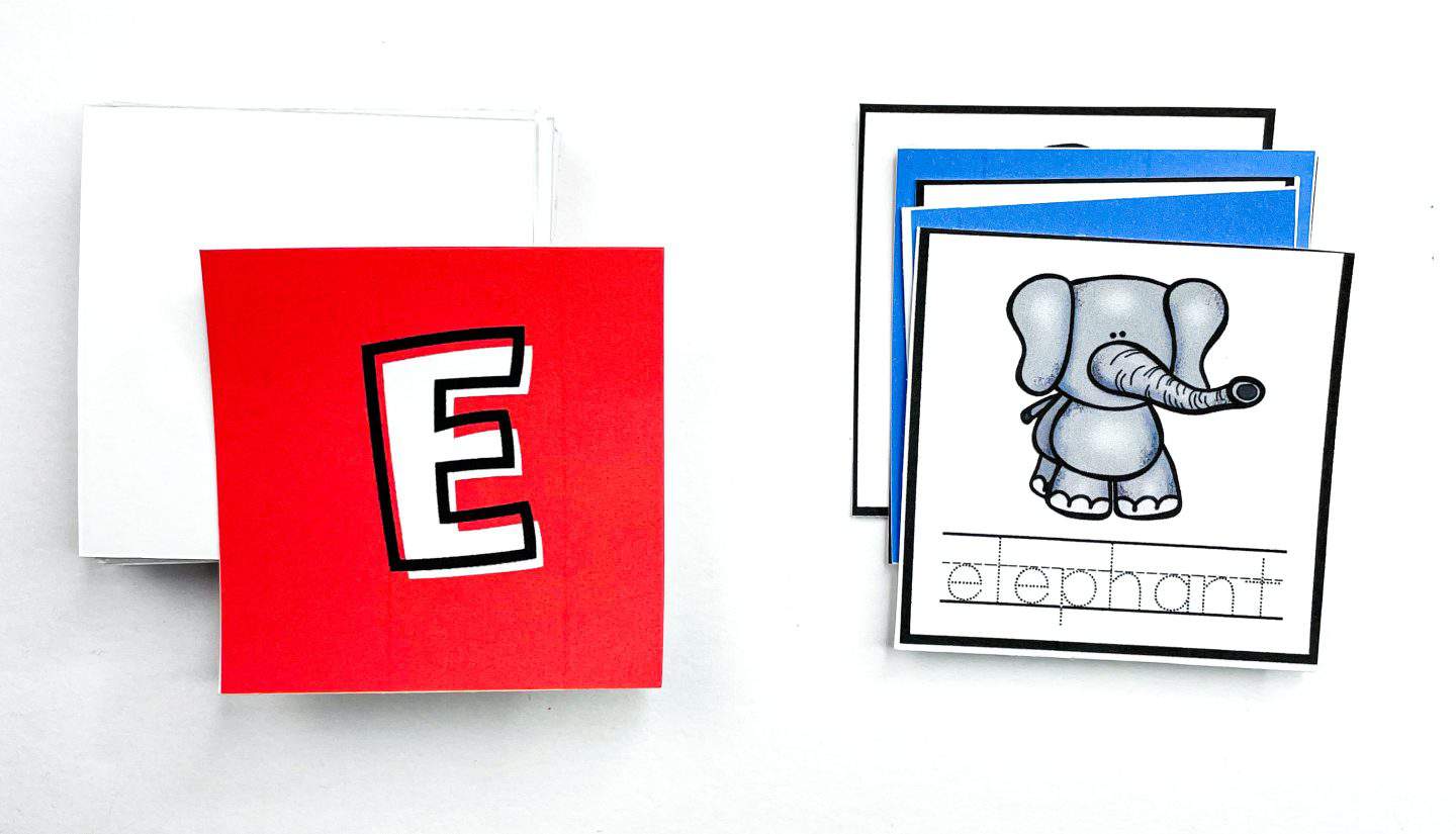 Alphabet matching game pieces showing a red card with the letter "E" and a corresponding card with a cute cartoon elephant labeled “elephant” in traceable handwriting. Additional blue letter cards are stacked in the background on a white surface.