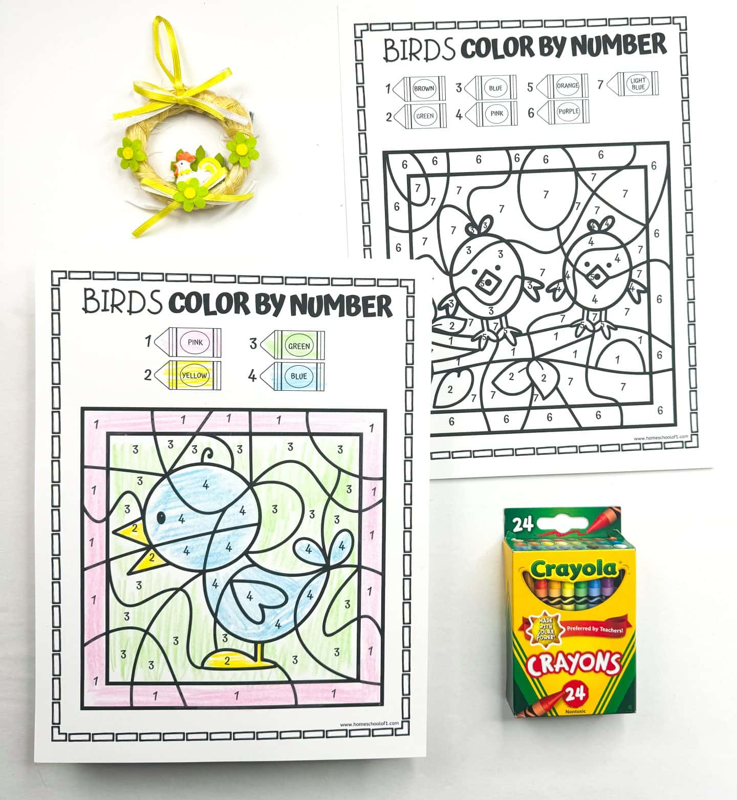 Two bird-themed color-by-number printable worksheets are displayed side by side. The completed sheet shows a blue bird with yellow feet against a pastel pink and green background, colored using pink, yellow, green, and blue crayons. A yellow decorative mini wreath with flowers and a Crayola 24-pack of crayons are also included in the layout.

