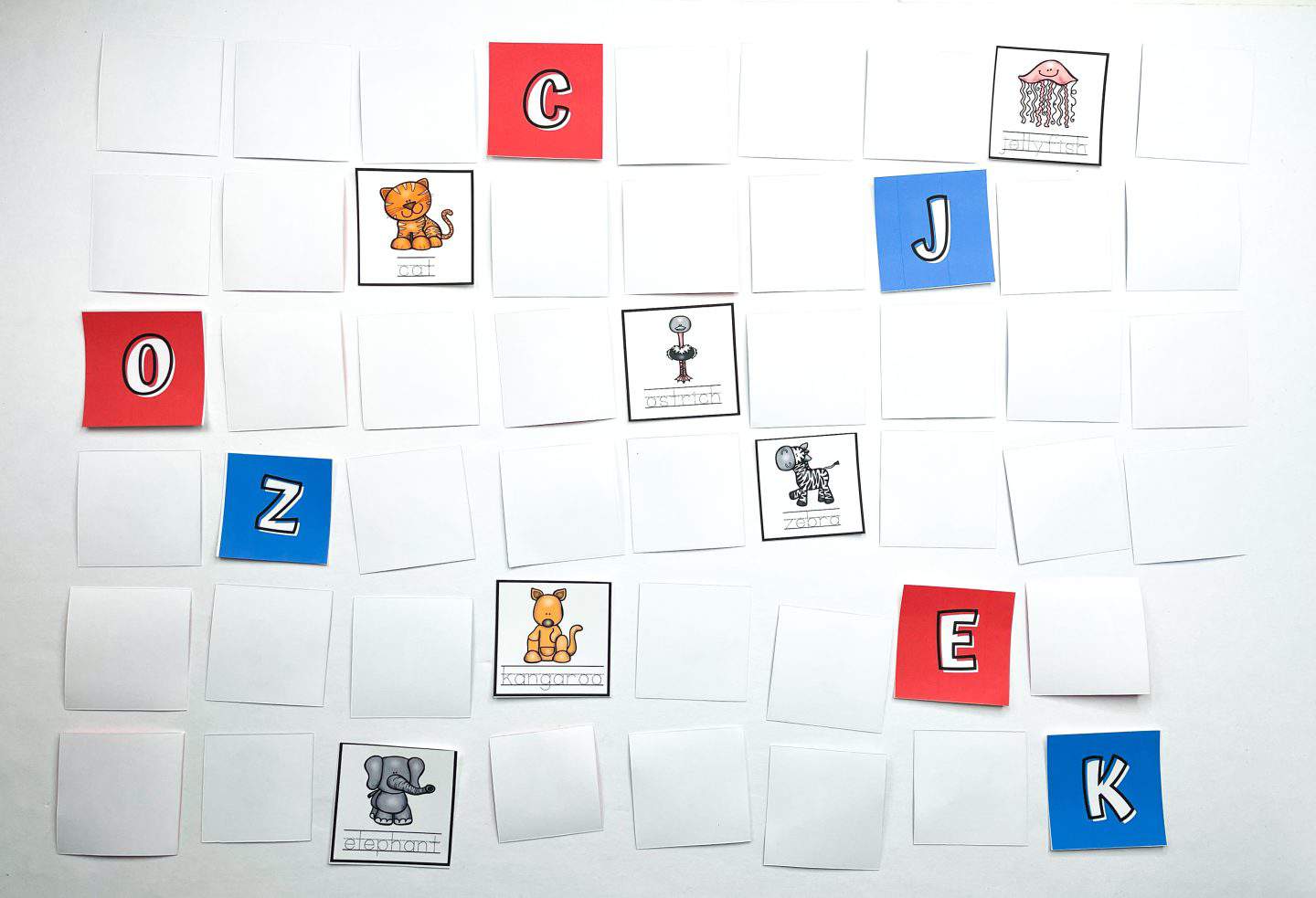 Alphabet matching game in progress, with some cards flipped over to reveal cartoon animal illustrations and corresponding words like cat, ostrich, jellyfish, zebra, kangaroo, and elephant. Red and blue letter cards are partially revealed, including the letters C, O, J, Z, E, and K. Cards are arranged in a grid on a white surface.