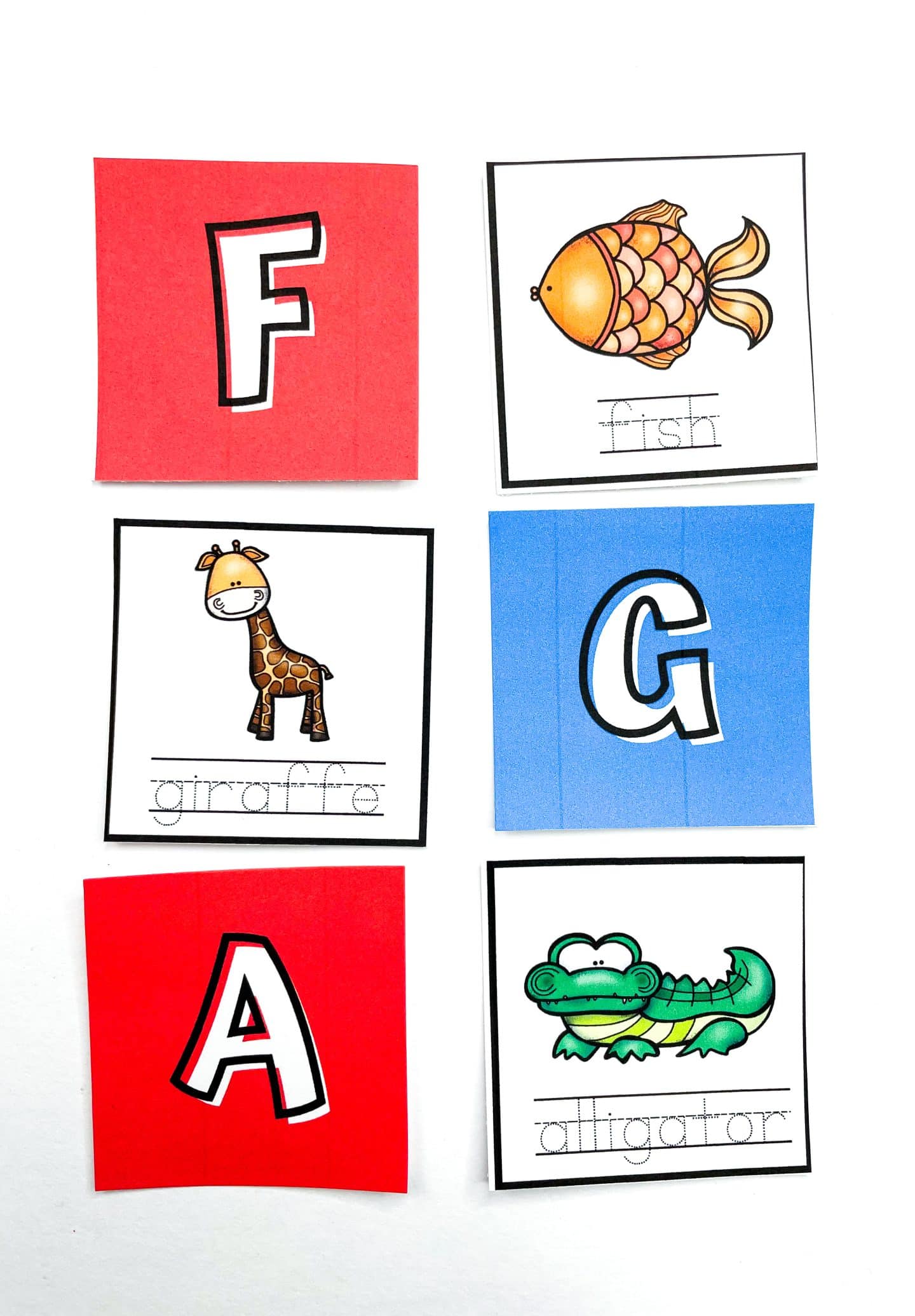 Close-up of an alphabet matching game showing three red and blue uppercase letter cards—F, G, and A—paired with illustrated animal cards labeled “fish,” “giraffe,” and “alligator.” Each animal card includes a cute cartoon drawing and a traceable word beneath it to support early literacy skills.