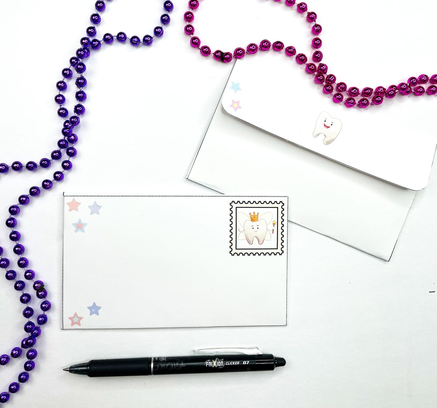 This image shows two finished Tooth Fairy envelopes—one sealed and one addressed—decorated with cute tooth graphics and colorful star designs. One envelope features a stamp illustration of a smiling tooth wearing a gold crown and holding a wand. A black Pilot FriXion Clicker pen and strings of purple and pink bead necklaces are also part of the scene, giving it a playful and festive vibe.