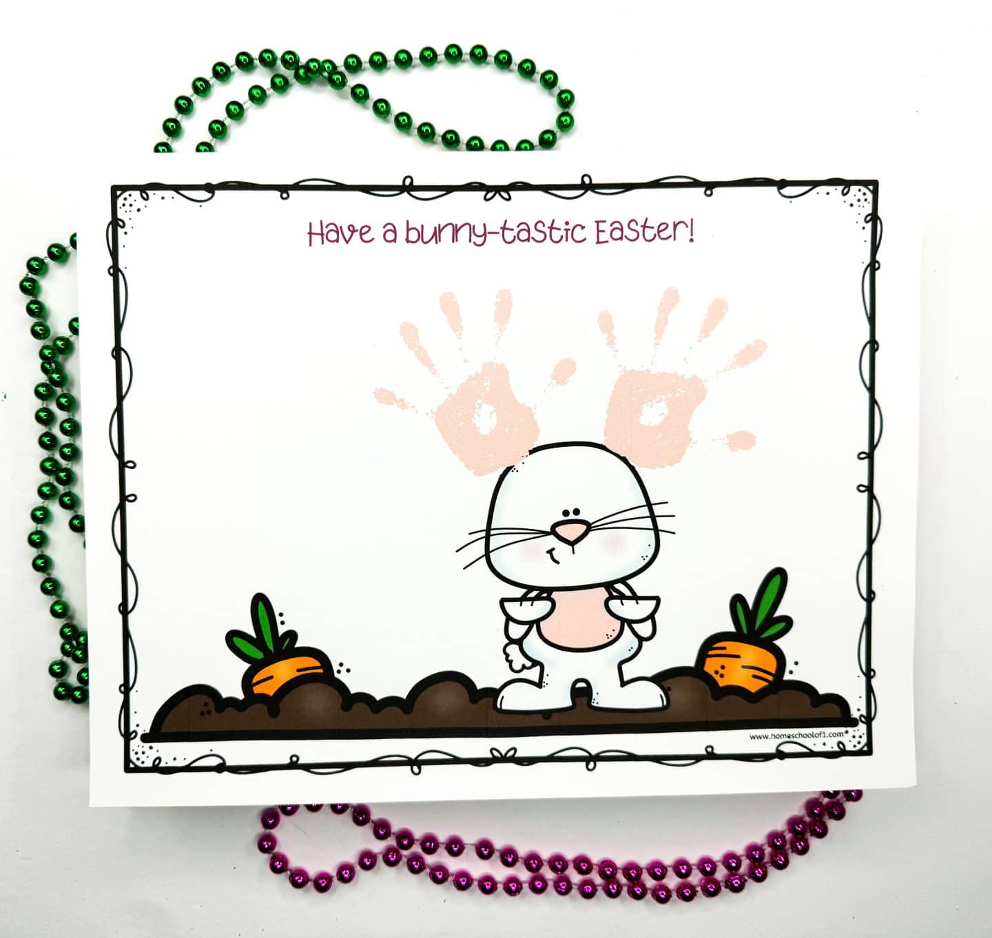 Completed Easter handprint art project with a bunny standing between two carrots and pink handprints added above its head to resemble bunny ears. The phrase “Have a bunny-tastic Easter!” is printed at the top in playful pink font. Green and purple beaded necklaces frame the cheerful craft, which is from homeschoolof1.com.