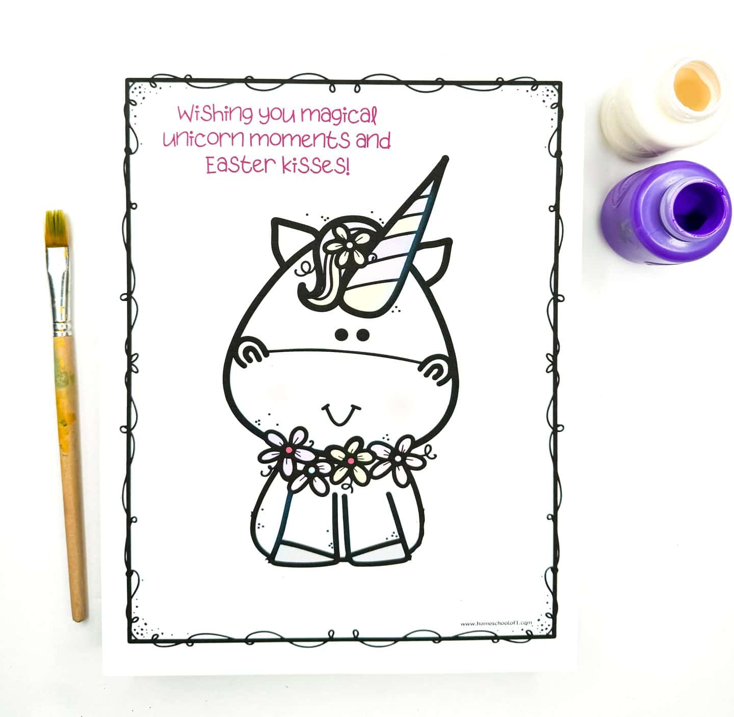Easter handprint art template featuring a unicorn with flowers and a rainbow-striped horn, surrounded by a whimsical border. The top reads “Wishing you magical unicorn moments and Easter kisses!” in pink text. Purple and cream paint bottles and a paintbrush are placed beside the paper.
