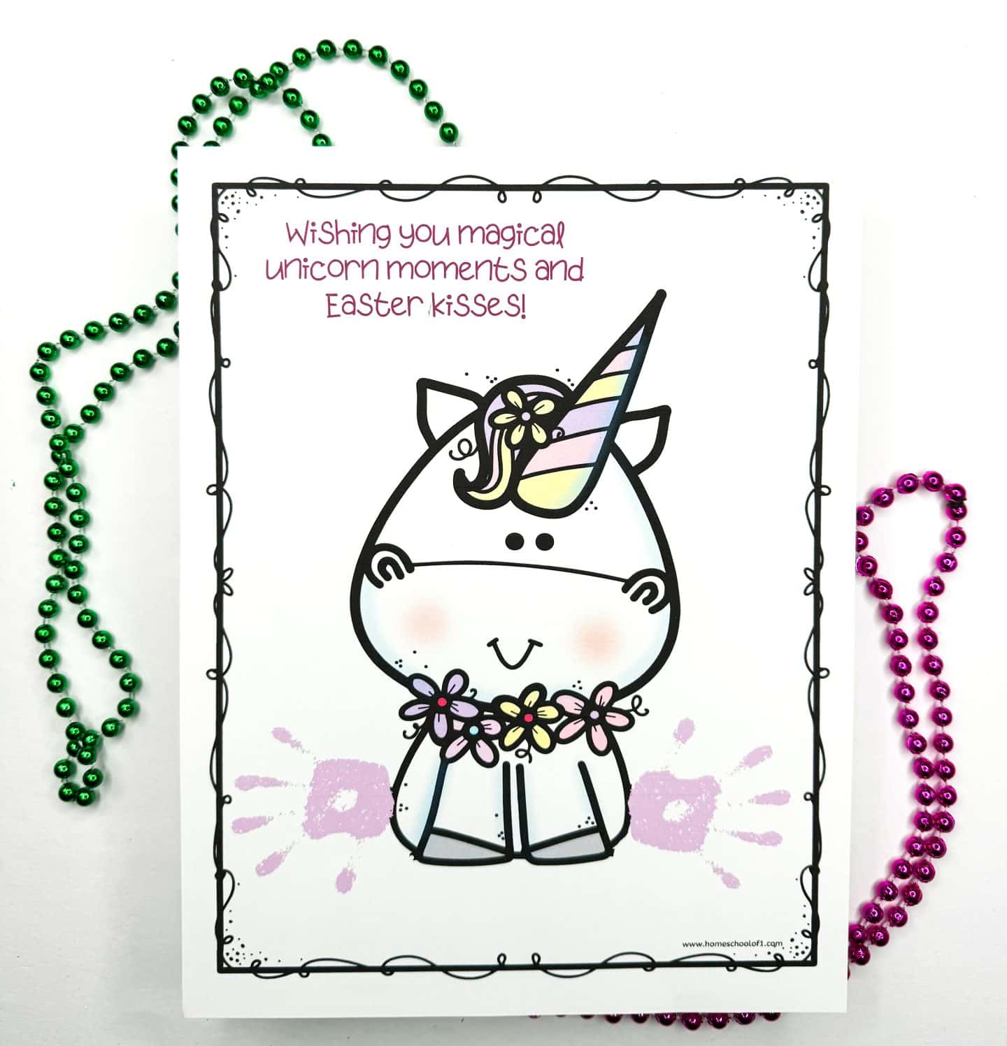 Completed Easter handprint art featuring a unicorn with a rainbow-striped horn and floral necklace. Two pastel purple handprints are stamped on either side of the unicorn’s lower body, creating a whimsical effect. The message at the top says, “Wishing you magical unicorn moments and Easter kisses!” Surrounded by green and pink bead necklaces.