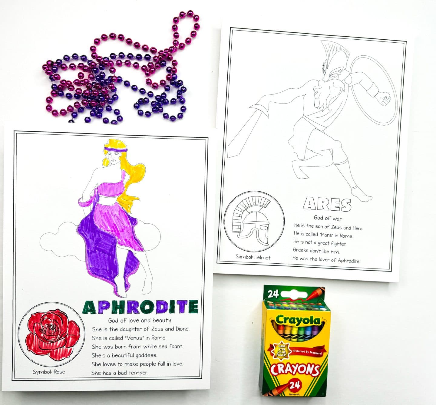 a colored-in Aphrodite, Goddess of love and beauty, in a purple dress with a rose symbol, and an uncolored Ares, God of war, holding a sword and shield. Purple and pink beads are scattered above the pages, and a Crayola crayon box is positioned at the bottom.