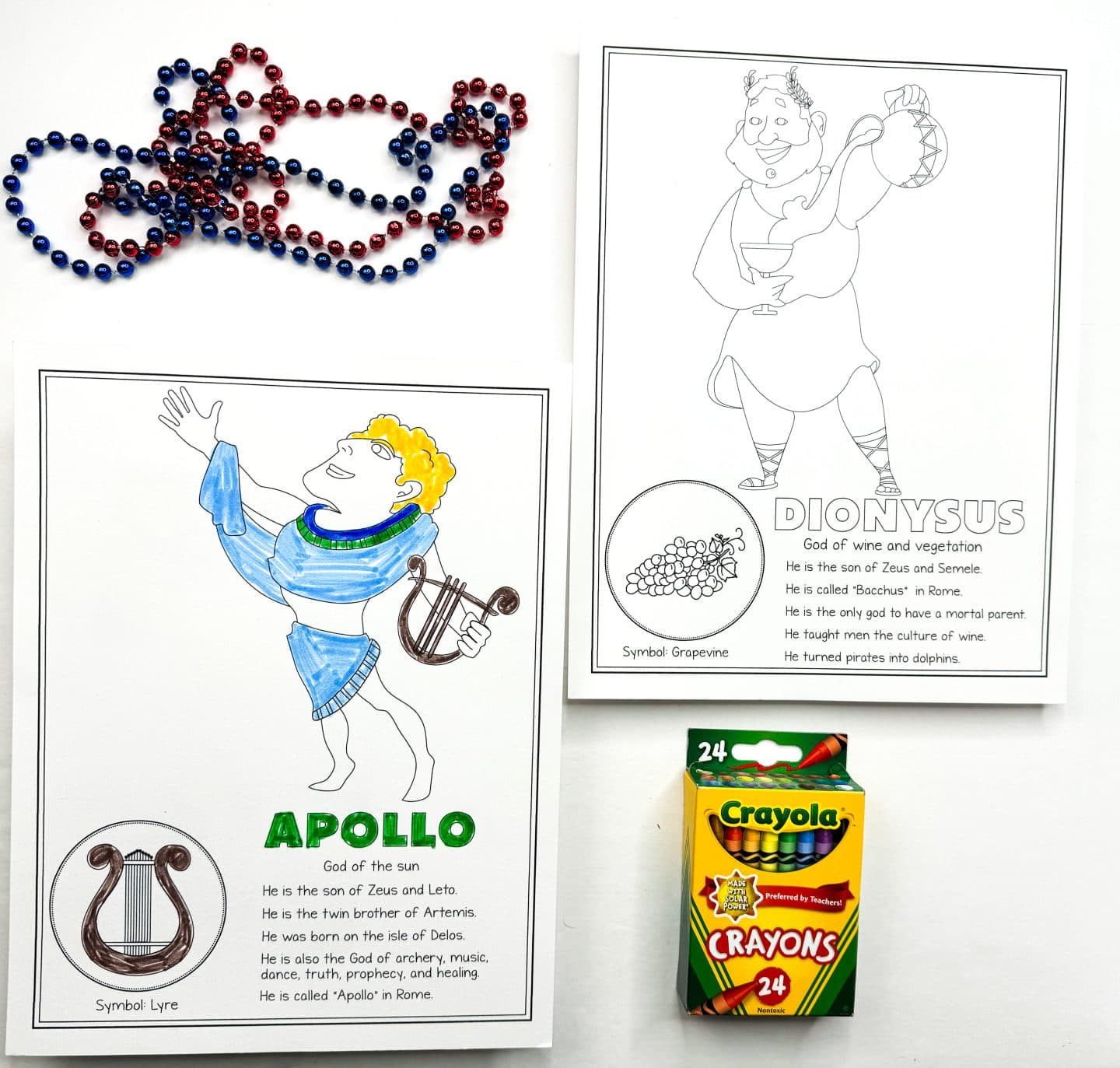 a colored-in Apollo, God of the sun, holding a lyre, with a blue and green outfit. Next to it is an uncolored page of Dionysus, God of wine and vegetation, holding a wine cup and a jar. Scattered red and blue beads are in the lower part of the image, and a Crayola crayon box is placed in the frame.