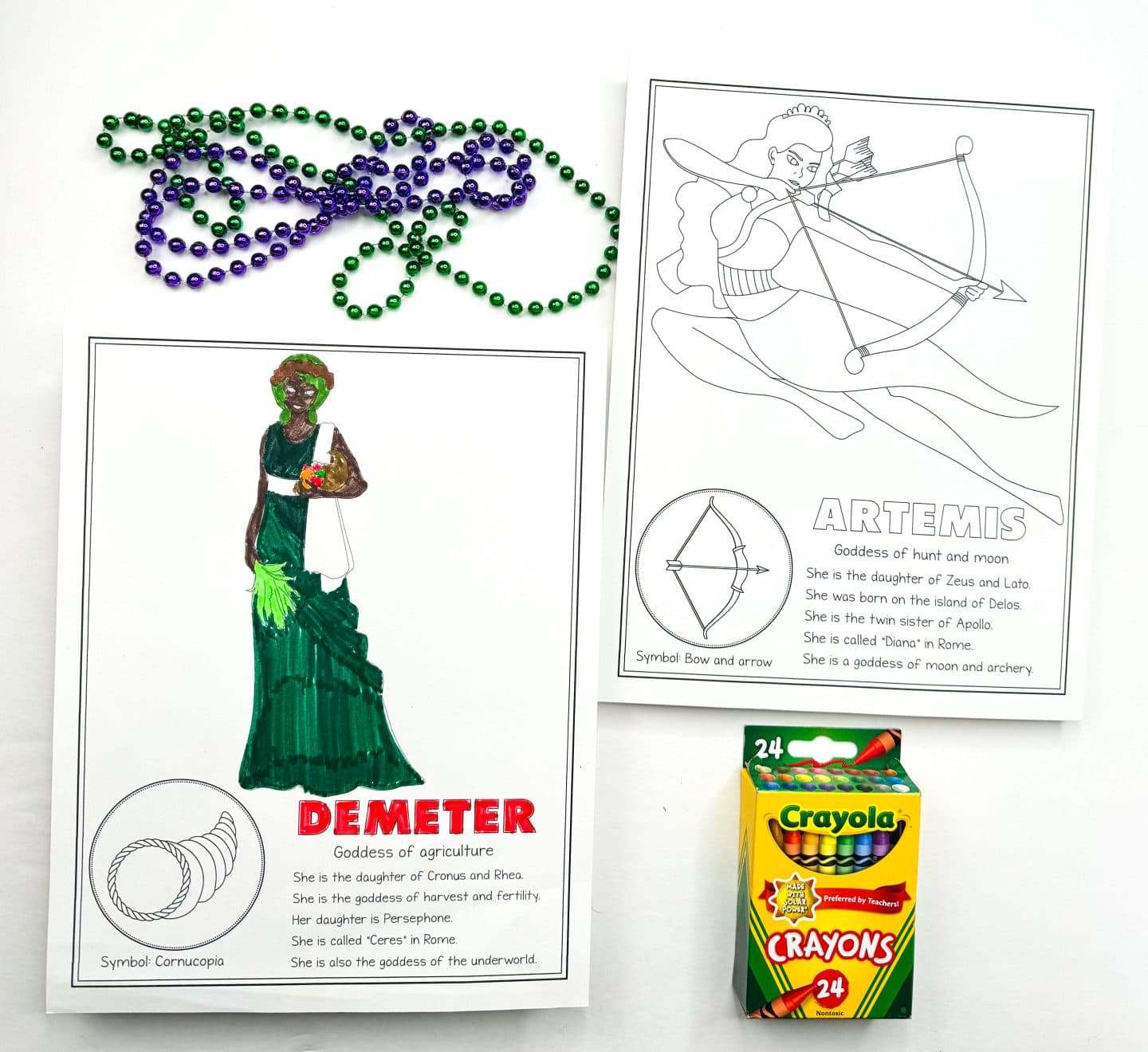  a colored-in Demeter, Goddess of agriculture, holding a cornucopia. Next to it is an uncolored page of Artemis, Goddess of hunt and moon, with a bow and arrow. There are green and purple beads scattered at the top and a Crayola crayon box placed in the frame.