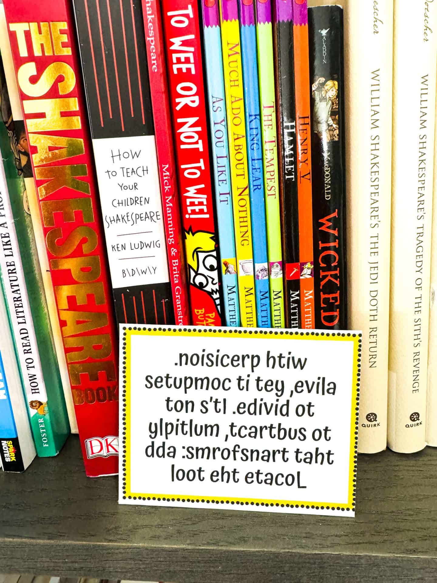 Easter egg hunt clue card placed on a bookshelf filled with Shakespeare-themed books. The clue is written in reversed text and reads: “Locate the tool that transforms: subtract, multiply, divide. It’s not alive, yet it computes with precision.” The setup suggests the next clue may be hidden near a calculator or computer. The card has a yellow dotted border and adds a clever, brain-teasing element to a teenage scavenger hunt.