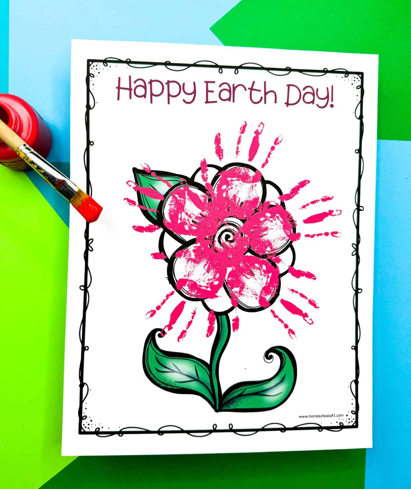 Earth Day flower handprint art featuring a large outlined flower with green leaves and bright pink painted handprints forming the petals. The page reads “Happy Earth Day!” in dark pink at the top, and it's surrounded by a whimsical border, placed on colorful green and blue paper with a red paintbrush and paint jar nearby.

