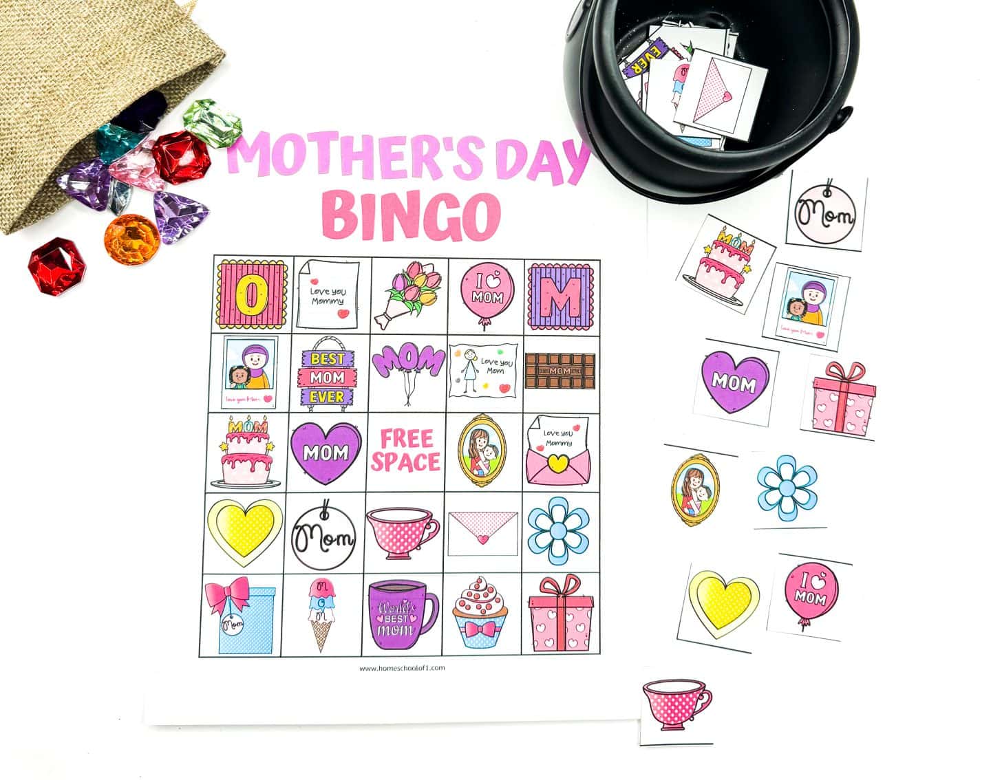 Mother’s Day Bingo game layout featuring a vibrant bingo card filled with themed illustrations like “Best Mom Ever,” cupcakes, flowers, teacups, gift boxes, and hearts with messages such as “Love you Mommy” and “I ? MOM.” Colorful plastic gem markers spill from a burlap pouch, and extra bingo tiles are spread around a black container to the right.

