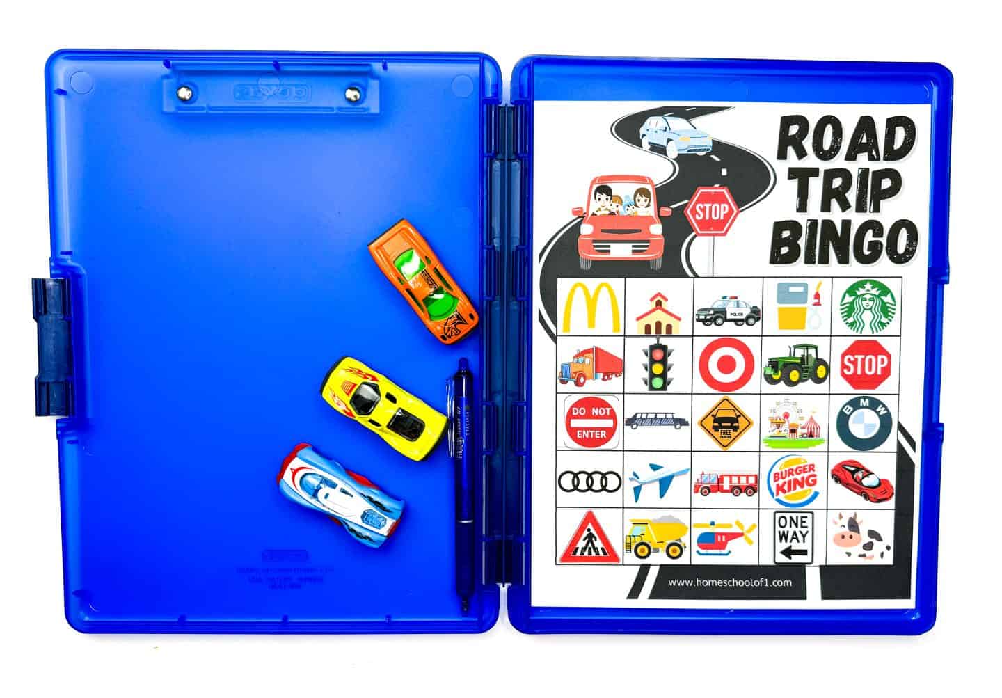 A laminated Road Trip Bingo card is displayed inside a bright blue clipboard case. The bingo grid features icons like McDonald's, Starbucks, police cars, traffic lights, gas stations, and various road signs. To the left of the case, three colorful toy race cars and a pen are positioned neatly, suggesting a fun travel activity for kids.