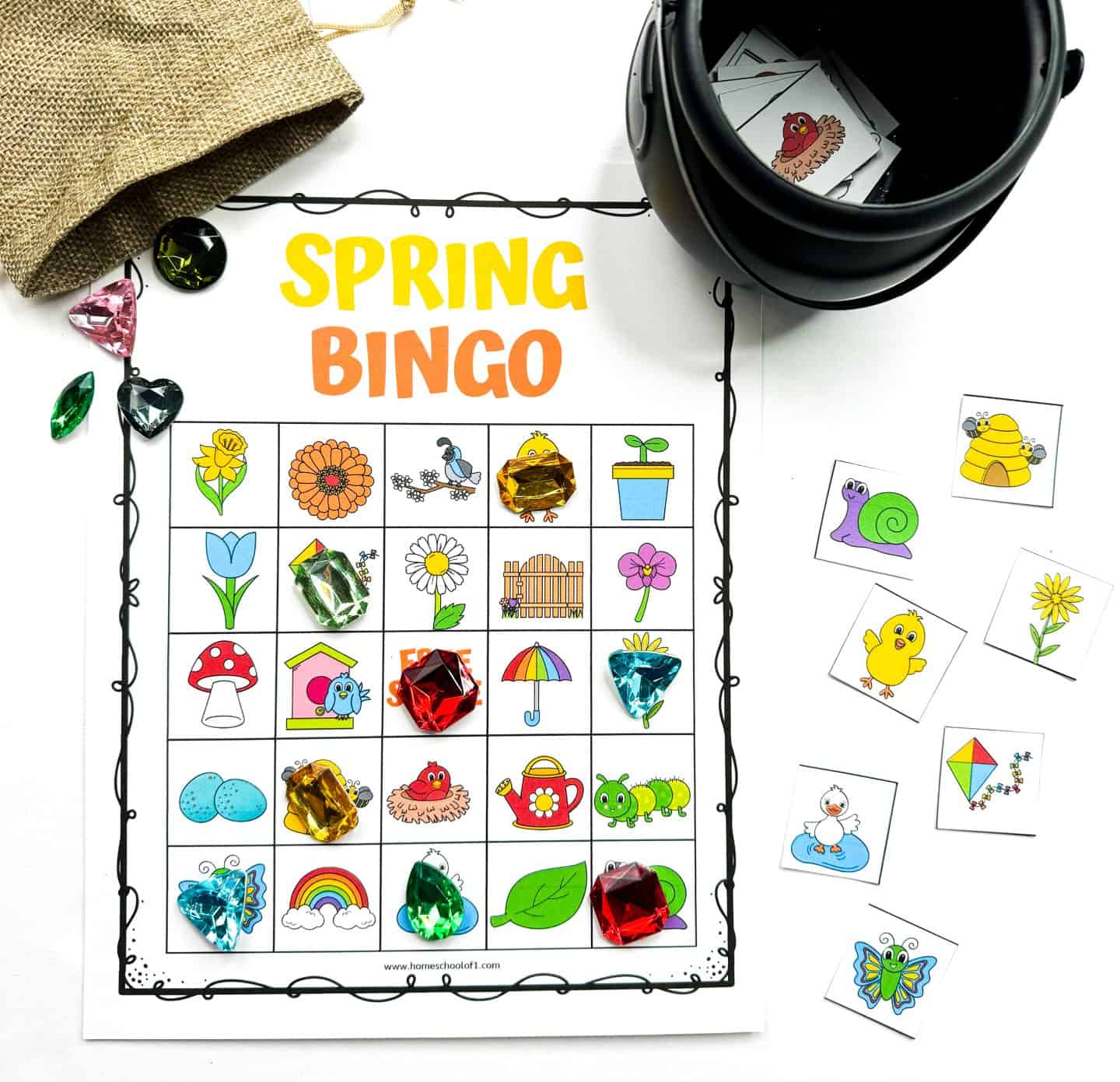 A completed spring-themed bingo card filled with colorful illustrations like flowers, rainbows, butterflies, and garden tools. Large gem markers highlight selected squares, with calling cards scattered nearby and a small black container and burlap pouch adding a playful touch to the seasonal kids' activity.