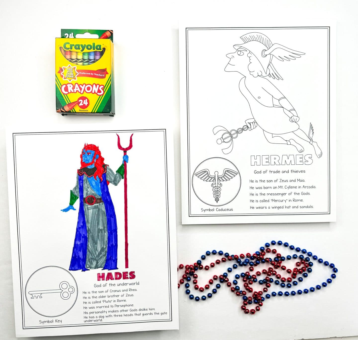 a colored-in Hades, God of the underworld, holding a trident. Next to it is an uncolored page of Hermes, God of trade and thieves, flying with his winged hat and sandals. There are red and blue beads scattered in the lower part of the image, and a Crayola crayon box is placed in the frame.