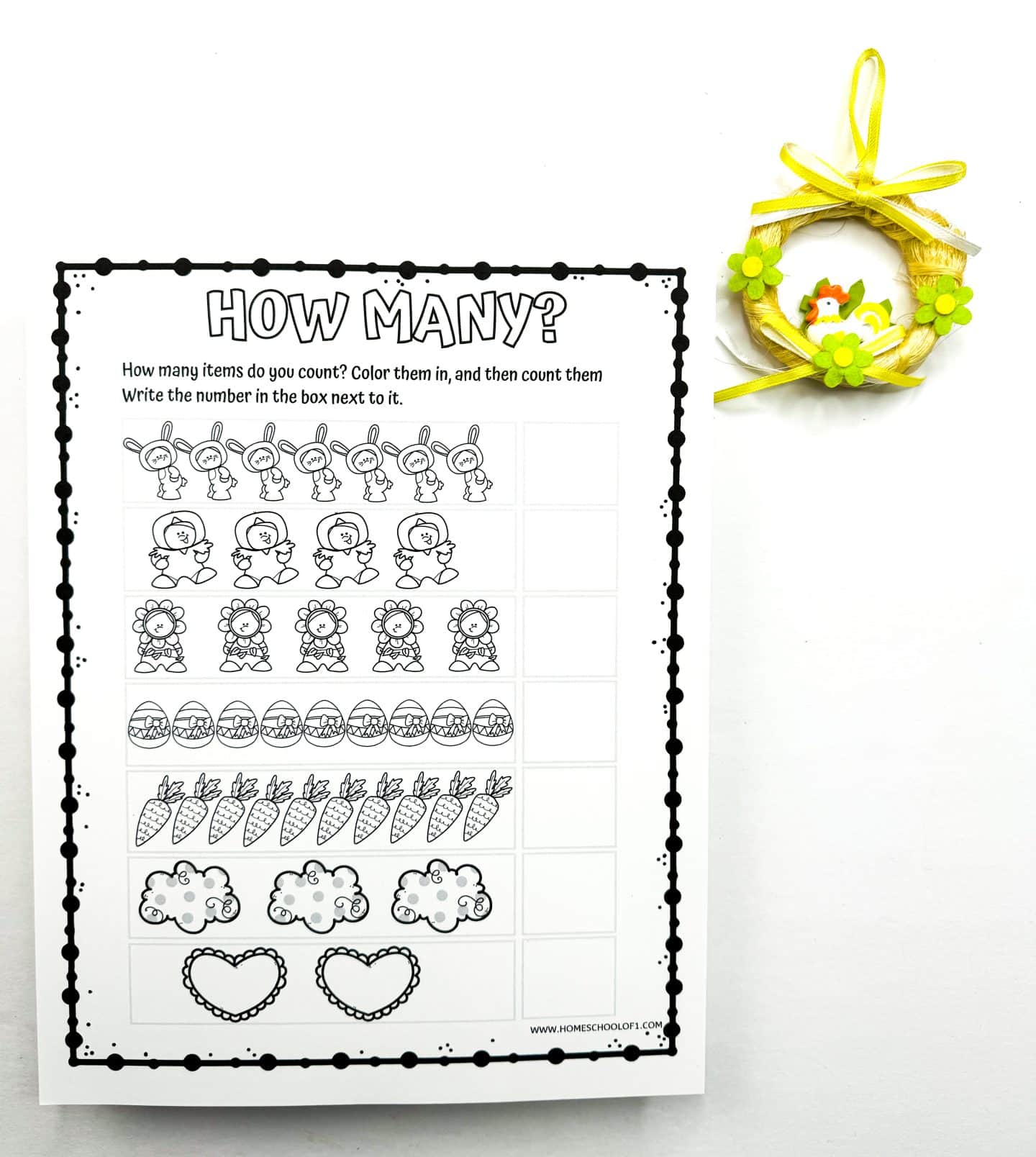  A black and white Easter-themed "How Many?" counting worksheet for kids featuring cute illustrations like bunnies, chicks, flowers, eggs, carrots, sheep, and hearts. Children are prompted to color the images, count each type, and write the total in the blank boxes beside them. A small yellow spring wreath with a chick decoration is placed in the corner.