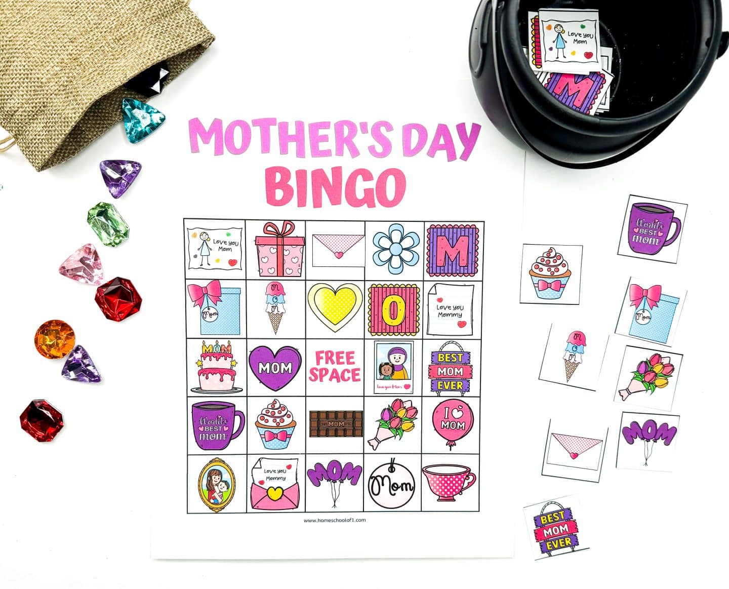 A bright and playful "Mother's Day Bingo" game with a colorful bingo card filled with themed illustrations such as hearts, cupcakes, flowers, and loving messages like "Love you Mom" and "Best Mom Ever." Decorative gem markers in jewel tones are scattered from a burlap pouch, and extra bingo tiles are placed to the right beside a black container.


