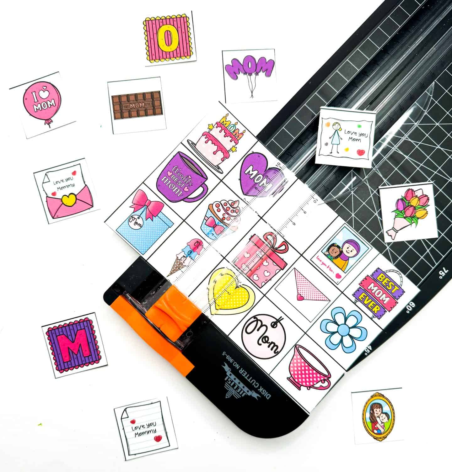 A paper trimmer is being used to cut out colorful Mother’s Day Bingo cards, each featuring themed illustrations like a cake, bouquet, chocolate bar, gift boxes, and hearts with messages such as “Love you Mom,” “I ? MOM,” and “Best Mom Ever.” Finished bingo squares are scattered around the trimmer, ready for gameplay or assembling.

