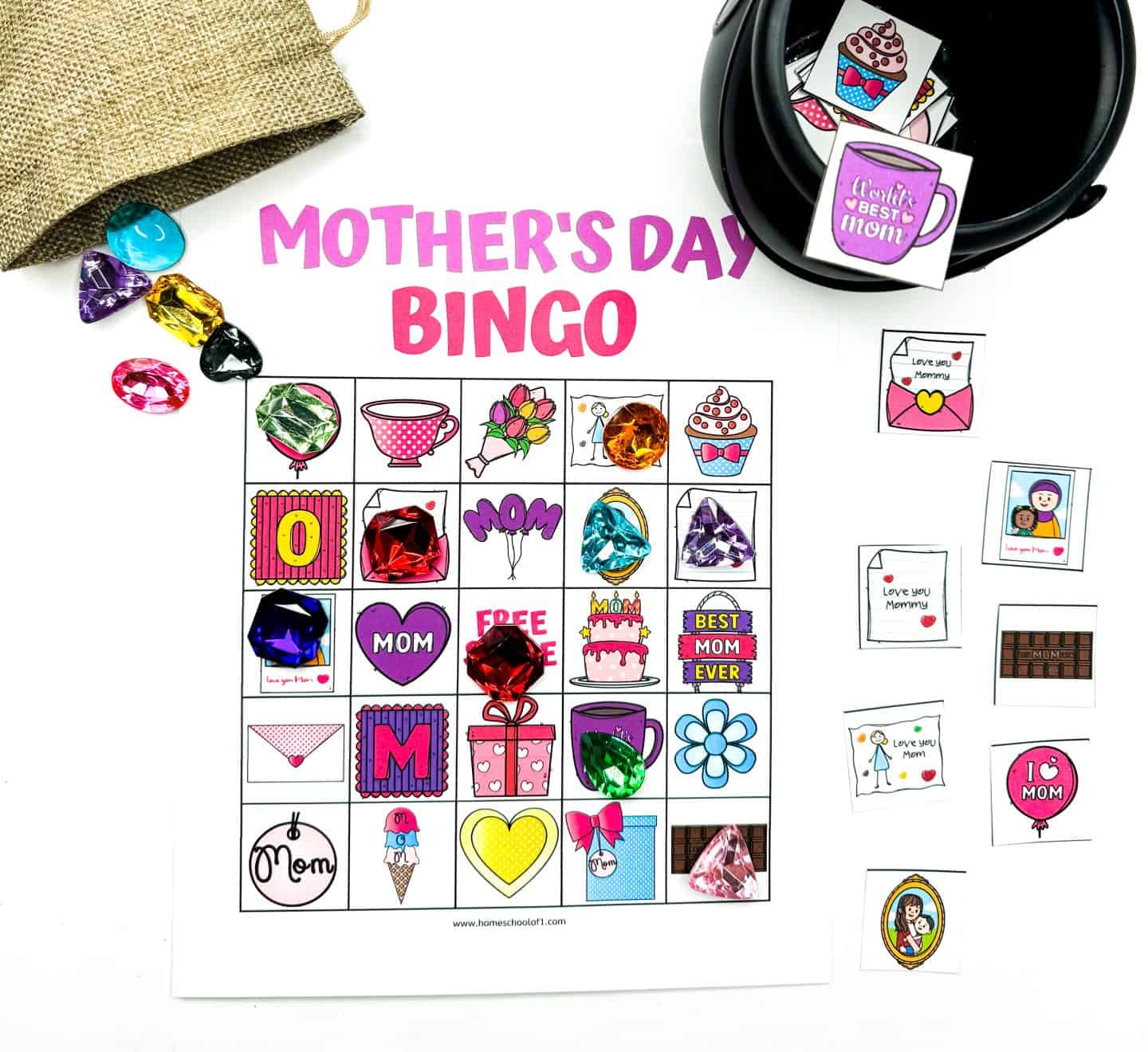 A colorful "Mother's Day Bingo" game set up with a printed bingo card featuring Mother's Day-themed illustrations like flowers, cupcakes, hearts, and messages like "Best Mom Ever." Large plastic gem markers in various colors are placed on some squares, with more spilling from a burlap pouch. Nearby, extra bingo cards and tokens with similar images and messages like "Love you Mommy" and "I ? MOM" are arranged.

