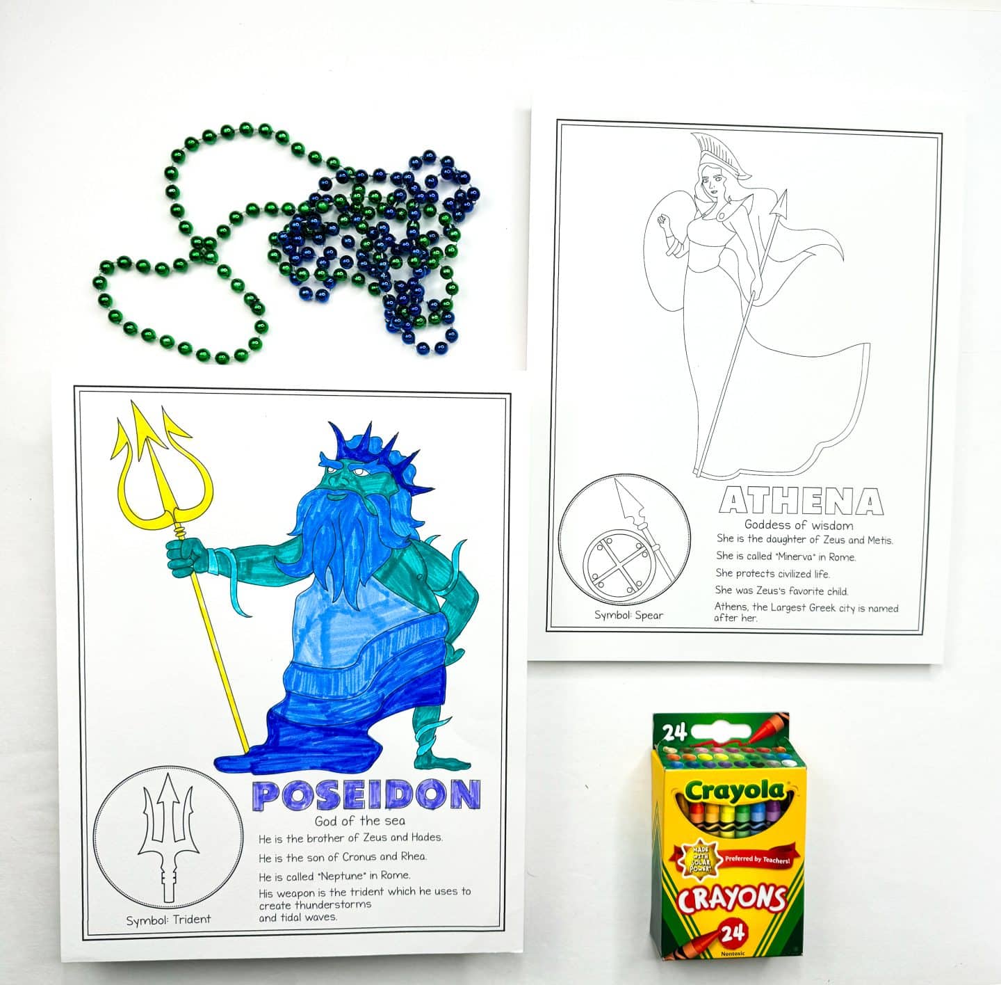 a colored-in Poseidon, the God of the sea, holding a trident. Next to it is an uncolored page of Athena, Goddess of wisdom. There are green and blue beads scattered at the top and a Crayola crayon box placed in the frame.