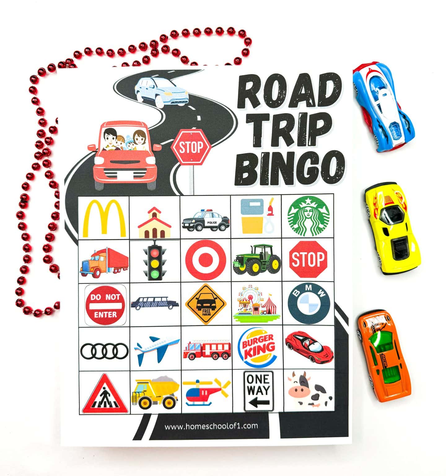 A printed “Road Trip Bingo” card featuring a variety of road-related icons such as McDonald's, Starbucks, stop signs, police cars, traffic lights, airplanes, and brand logos like Target, BMW, and Burger King. The sheet is surrounded by toy cars and a red bead necklace, making it look playful and travel-ready. The top of the page shows a family driving on a road with a stop sign illustration.
