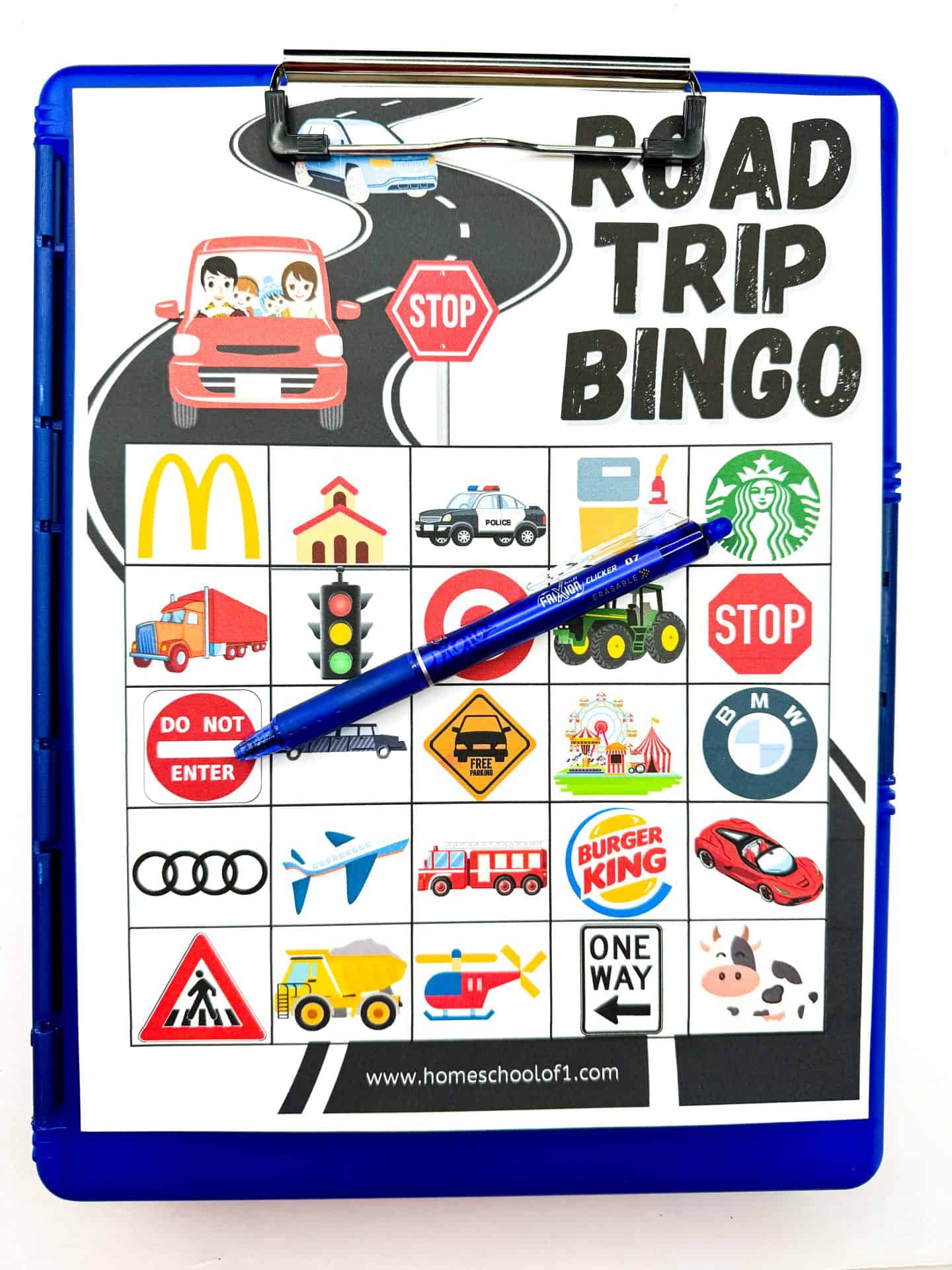 Colorful road trip bingo game sheet clipped to a blue clipboard with a pen resting on top. The bingo card includes icons such as McDonald's, Starbucks, a stop sign, traffic lights, a police car, an airplane, and various vehicles and road signs. The top of the sheet features the title "Road Trip Bingo" in bold black letters with an illustration of a family in a red car on a winding road.