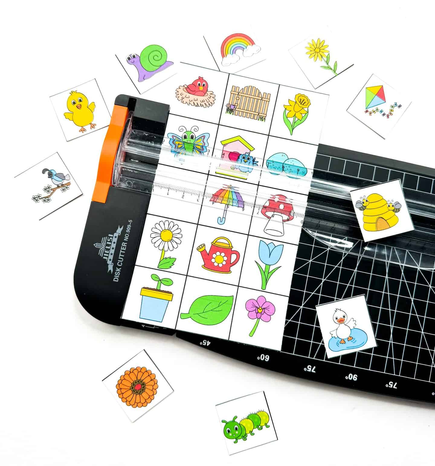 Spring-themed bingo calling cards being cut with a paper trimmer, featuring colorful illustrations such as a caterpillar, beehive, rainbow, daisy, watering can, and butterfly. These printable bingo pieces are part of a fun seasonal game for kids.
