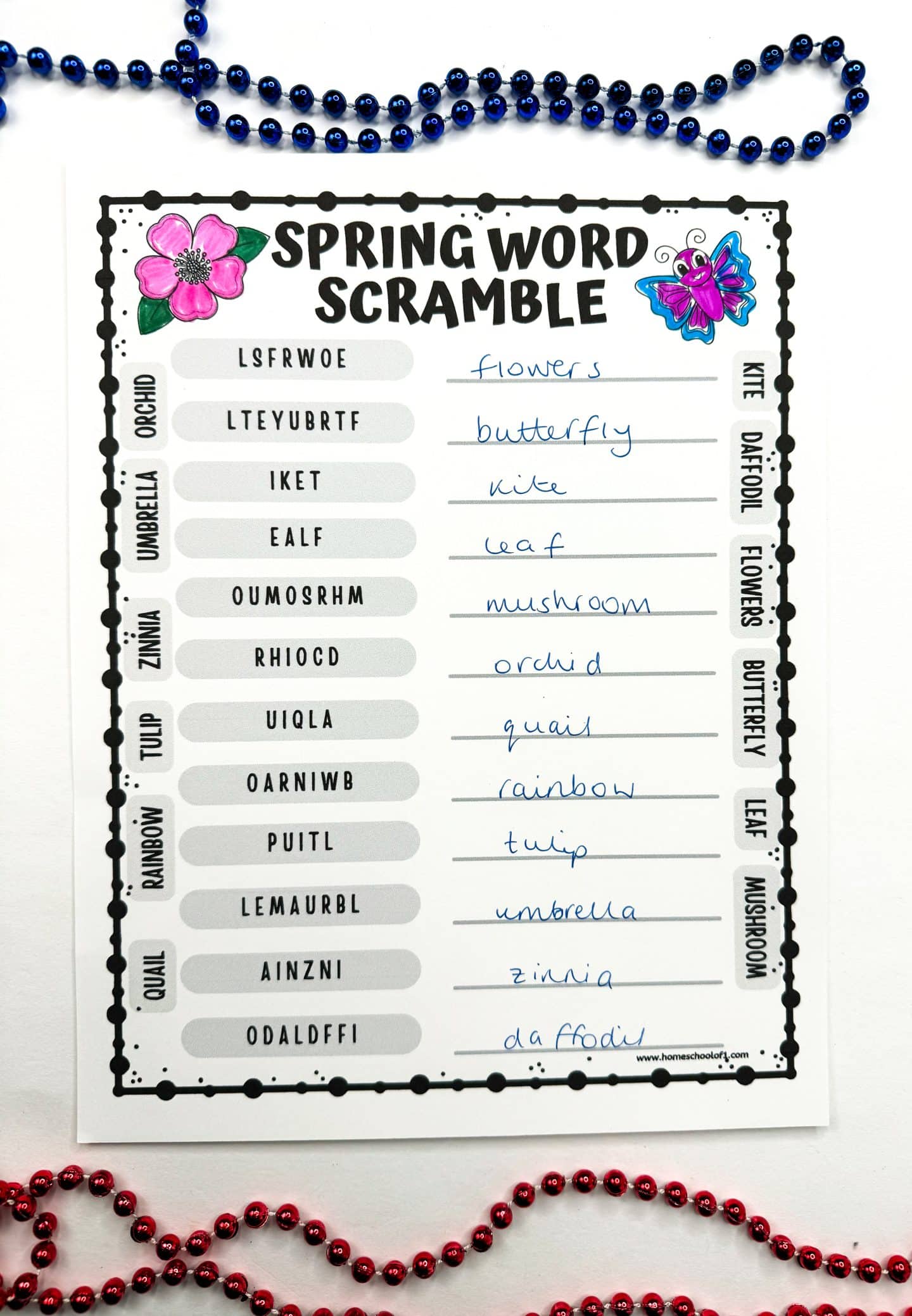 A completed Spring Word Scramble worksheet features hand-written answers like flowers, butterfly, kite, and mushroom next to scrambled words. The worksheet includes decorative spring elements like a pink flower and a cartoon butterfly, with a vertical word bank on the right. Blue and red beaded necklaces frame the page for a festive, springtime look.